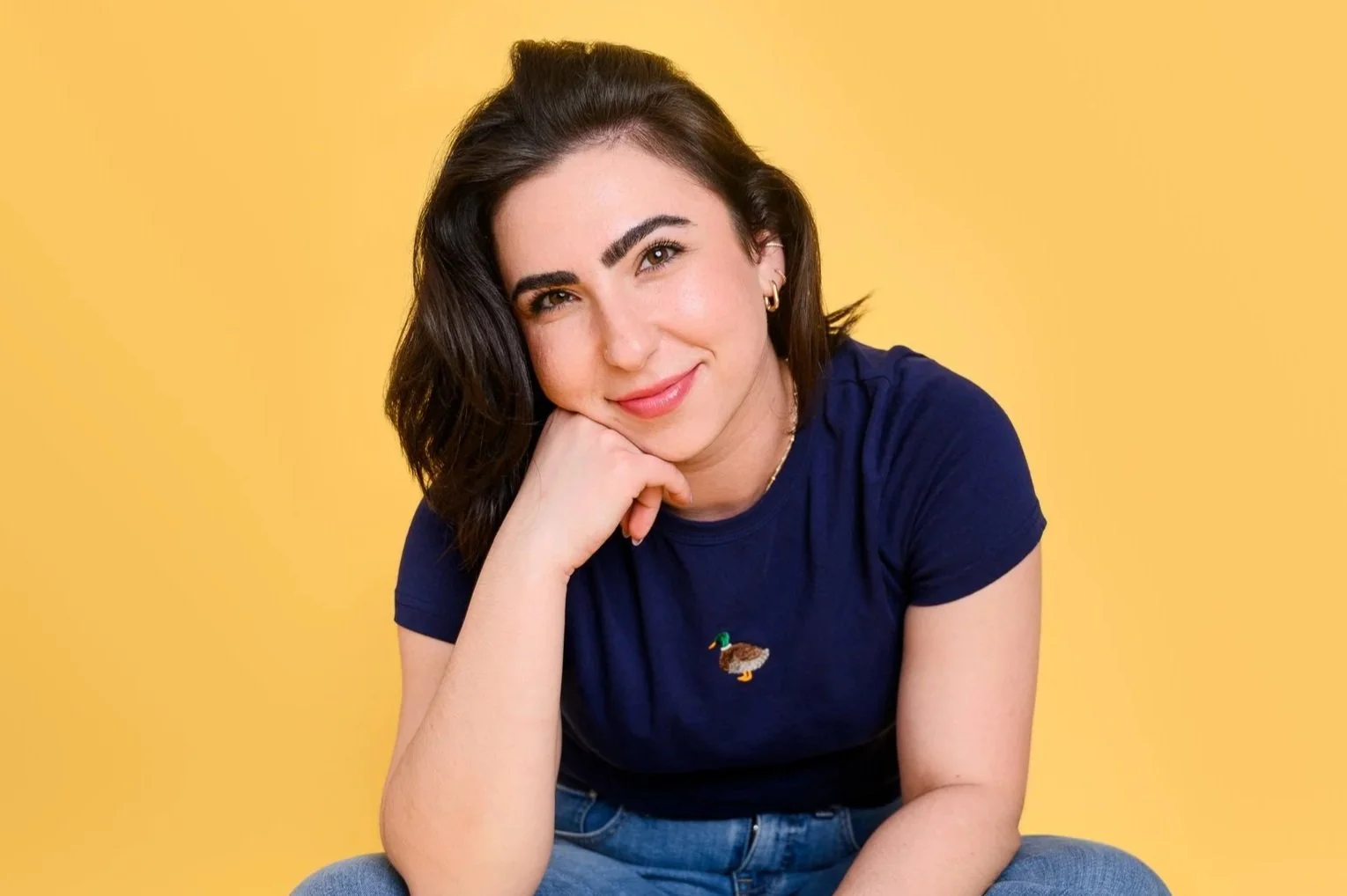Young woman with dark hair, wearing a navy blue t-shirt with a small embroidered duck and blue jeans, posing against a yellow background, resting her chin on her hand and smiling softly.