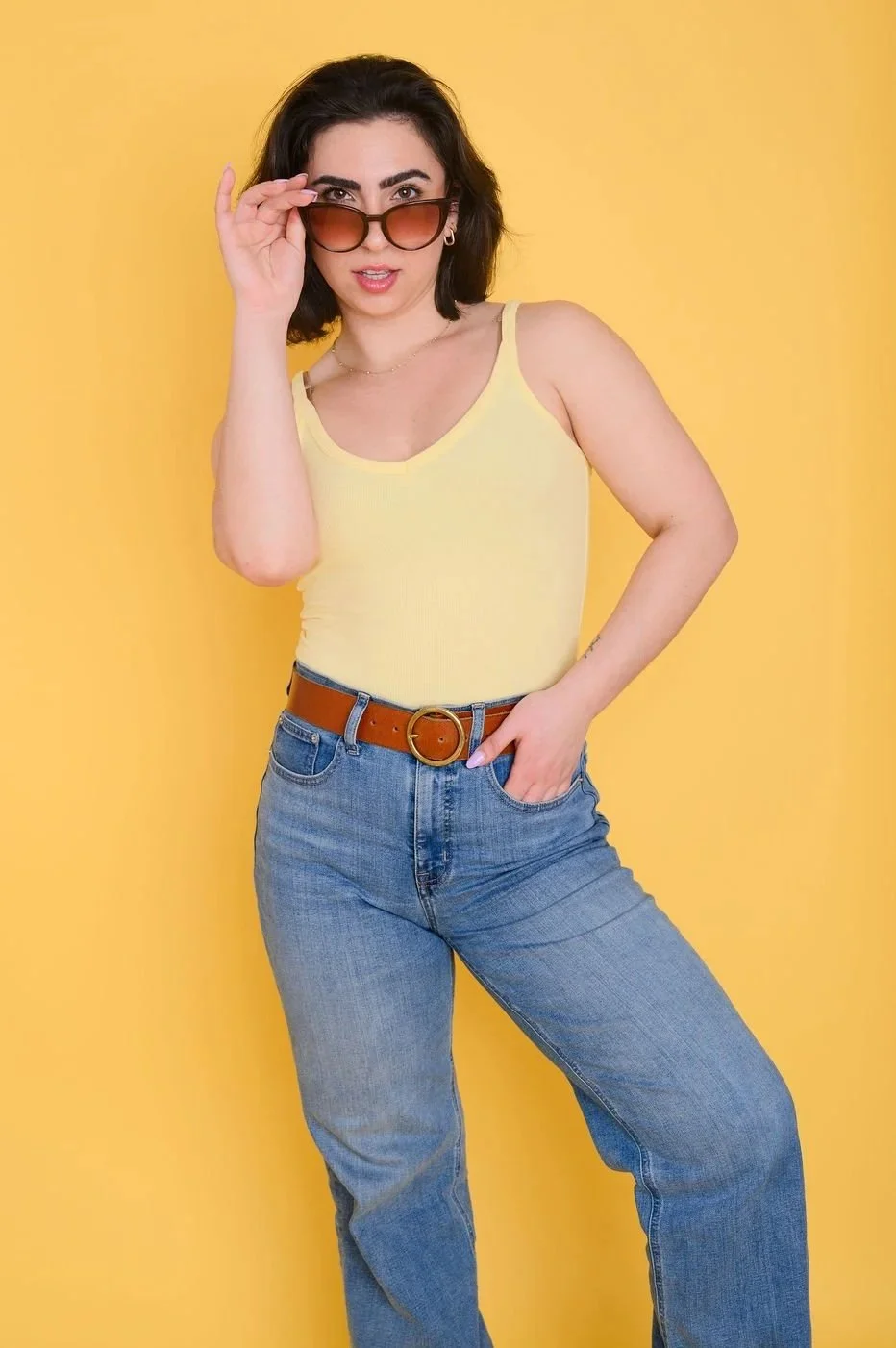 Woman with dark hair wearing a yellow tank top, blue jeans, a brown belt, and sunglasses against a yellow background.