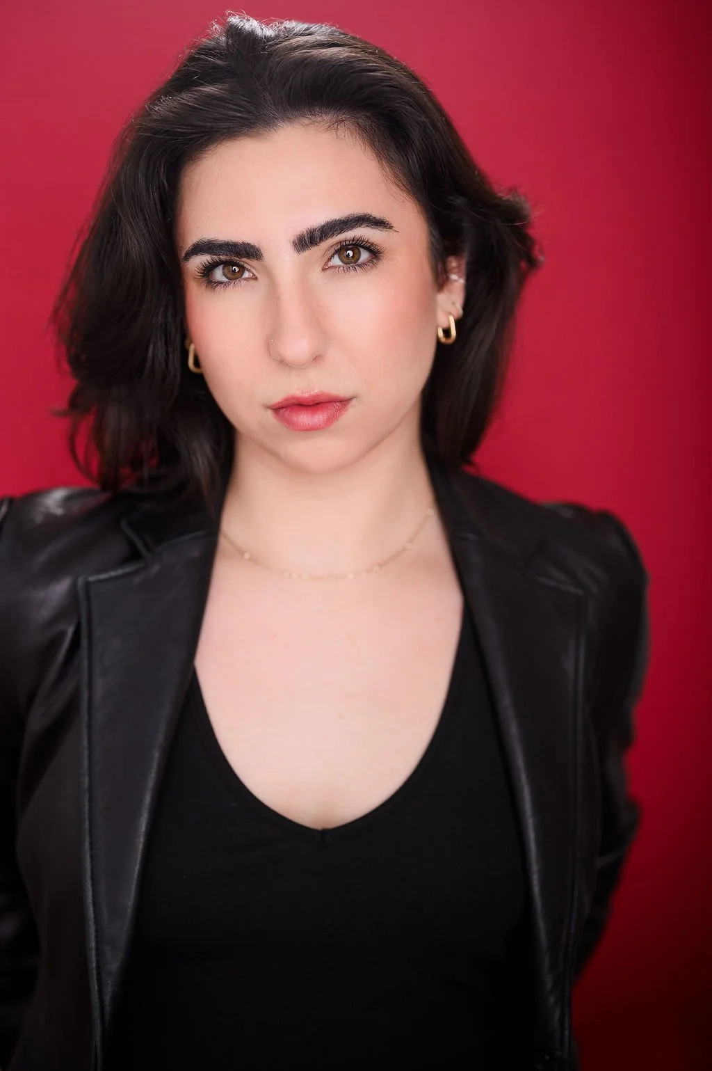 Portrait of a young woman with dark hair, wearing gold jewelry, a black top, and a leather jacket against a red background.
