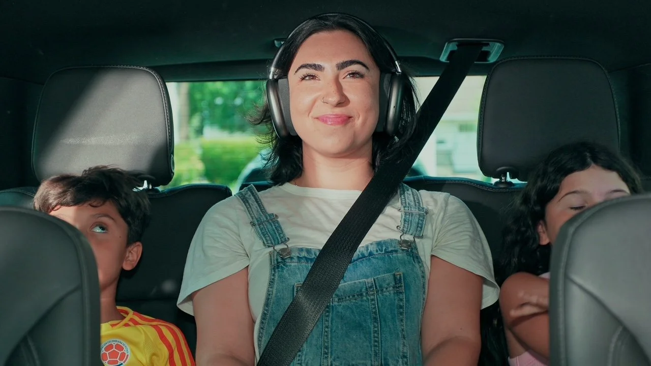 A woman with headphones smiling in the back seat of a car, with two children sleeping on either side of her.