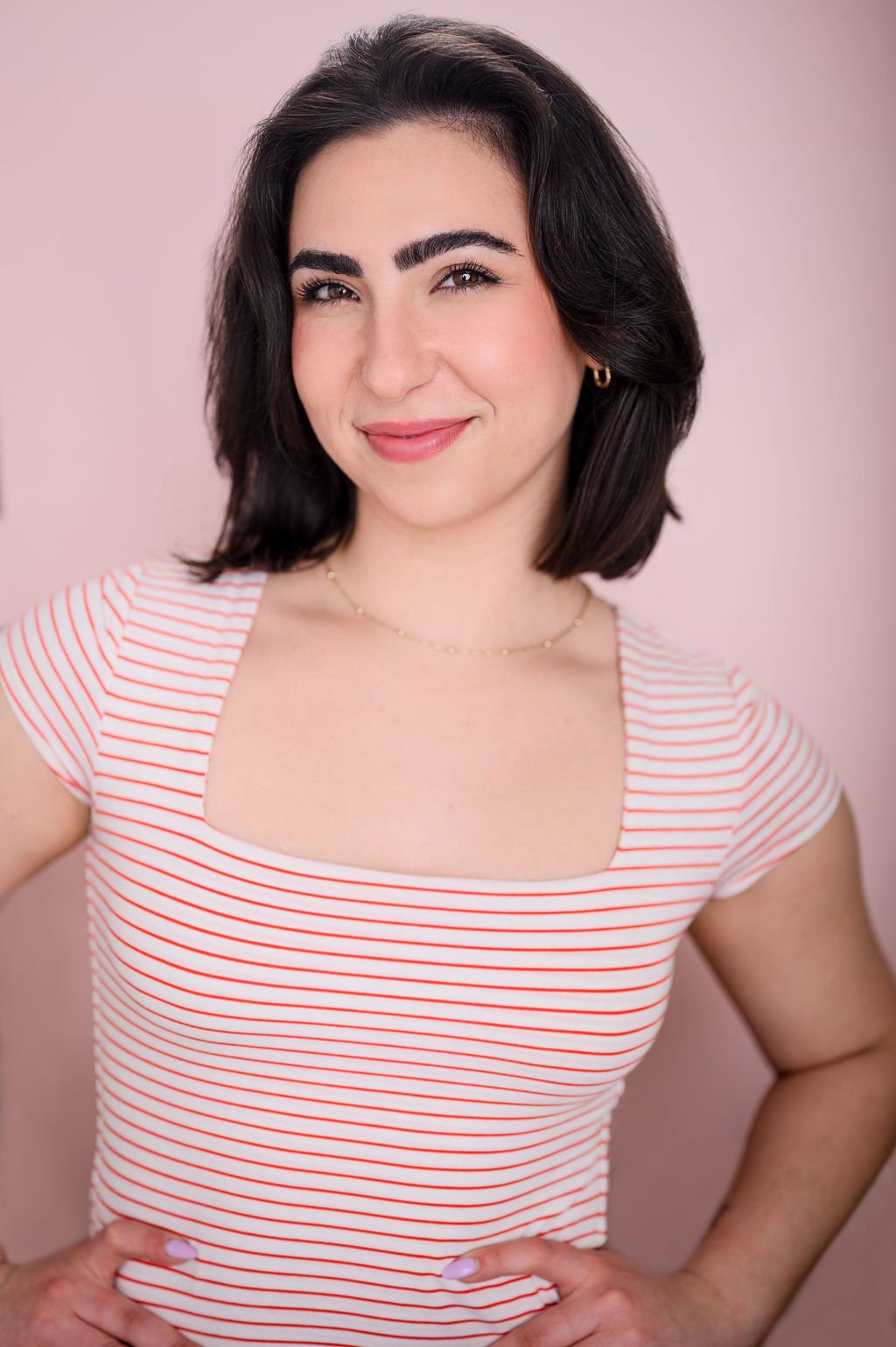 A young woman with dark, shoulder-length hair and light skin, smiling and looking at the camera against a light pink background. She is wearing a striped, short-sleeved, square-neck top and small gold earrings.
