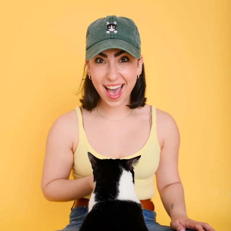 A young woman with dark hair, wearing a green cap, yellow tank top, and jewelry, is smiling excitedly while sitting against a yellow background, with a black and white cat sitting in front of her, facing away.