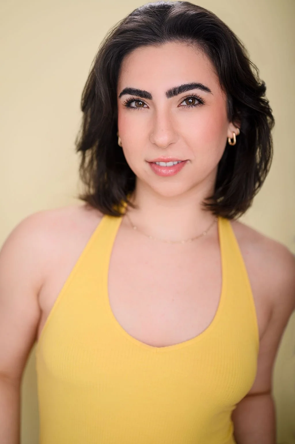 A young woman with shoulder-length dark hair and light skin wearing a yellow sleeveless top, earrings, and a delicate necklace, smiling at the camera against a plain light-colored background.