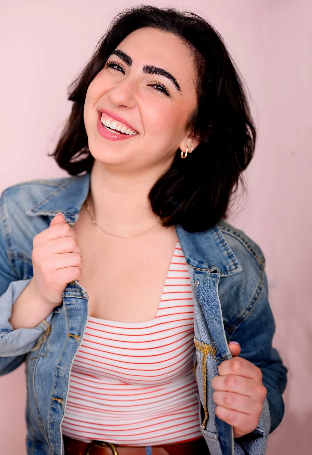 A woman with dark hair smiling, wearing a denim jacket and a red and white striped top, against a pink background.