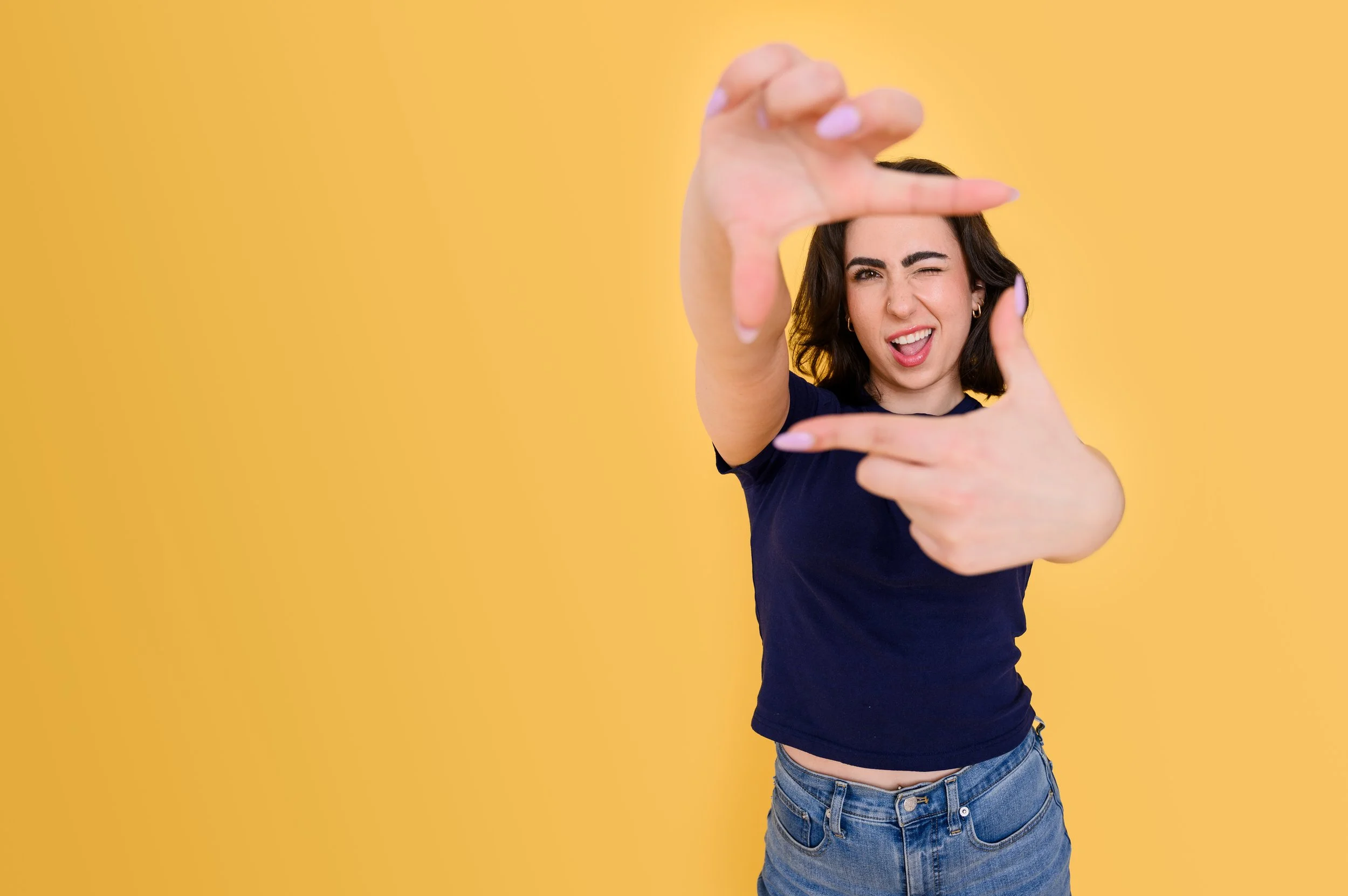 A woman wearing a navy T-shirt and jeans frames the camera with her hands and poses with one eye closed against a yellow background.