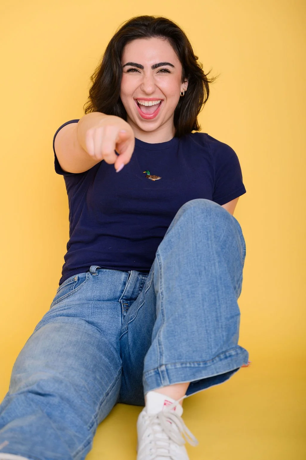 A smiling young woman with dark hair, wearing a navy blue t-shirt with a duck embroidered on it, blue jeans, and white sneakers, sitting against a yellow background, pointing at the camera.