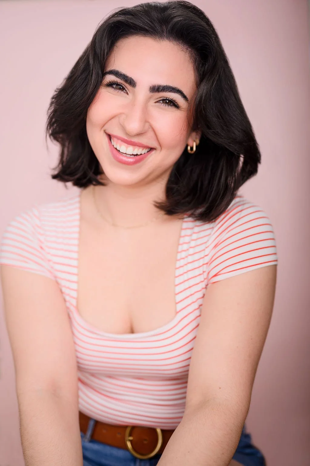 A young woman with dark wavy hair, smiling and wearing a striped pink and white top, earrings, and a belt, against a pink background.