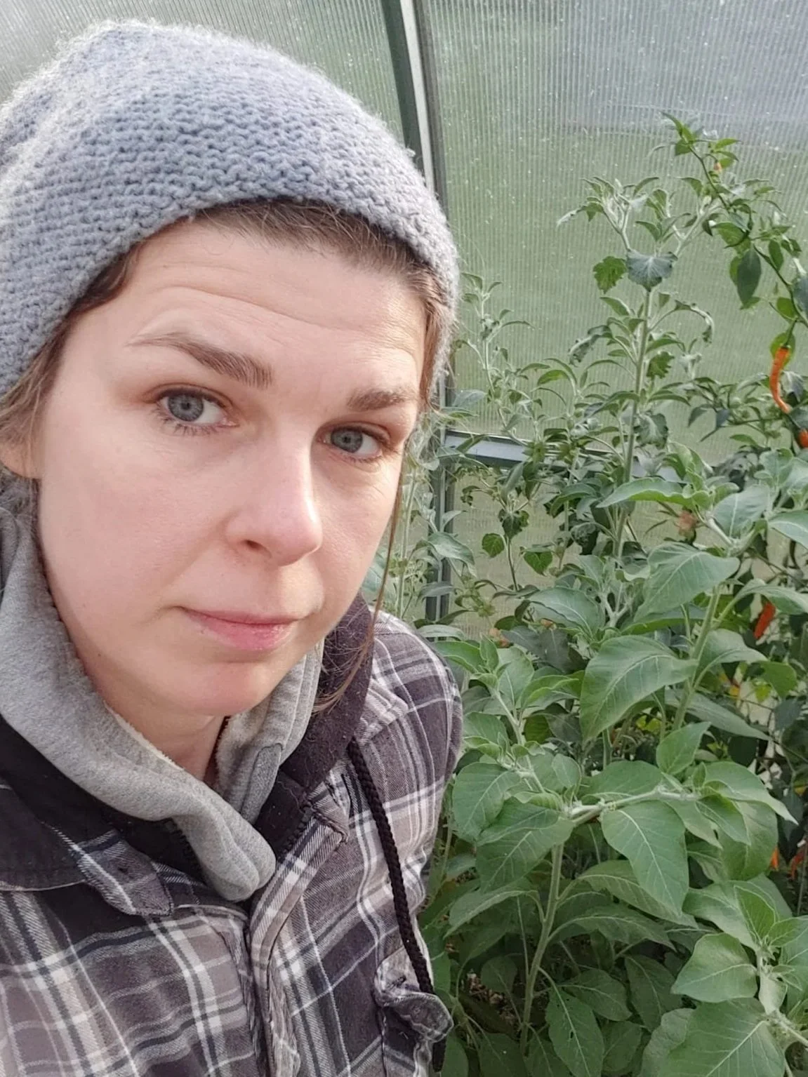 A woman in a gray beanie and plaid jacket taking a selfie in a greenhouse with green plants around her.