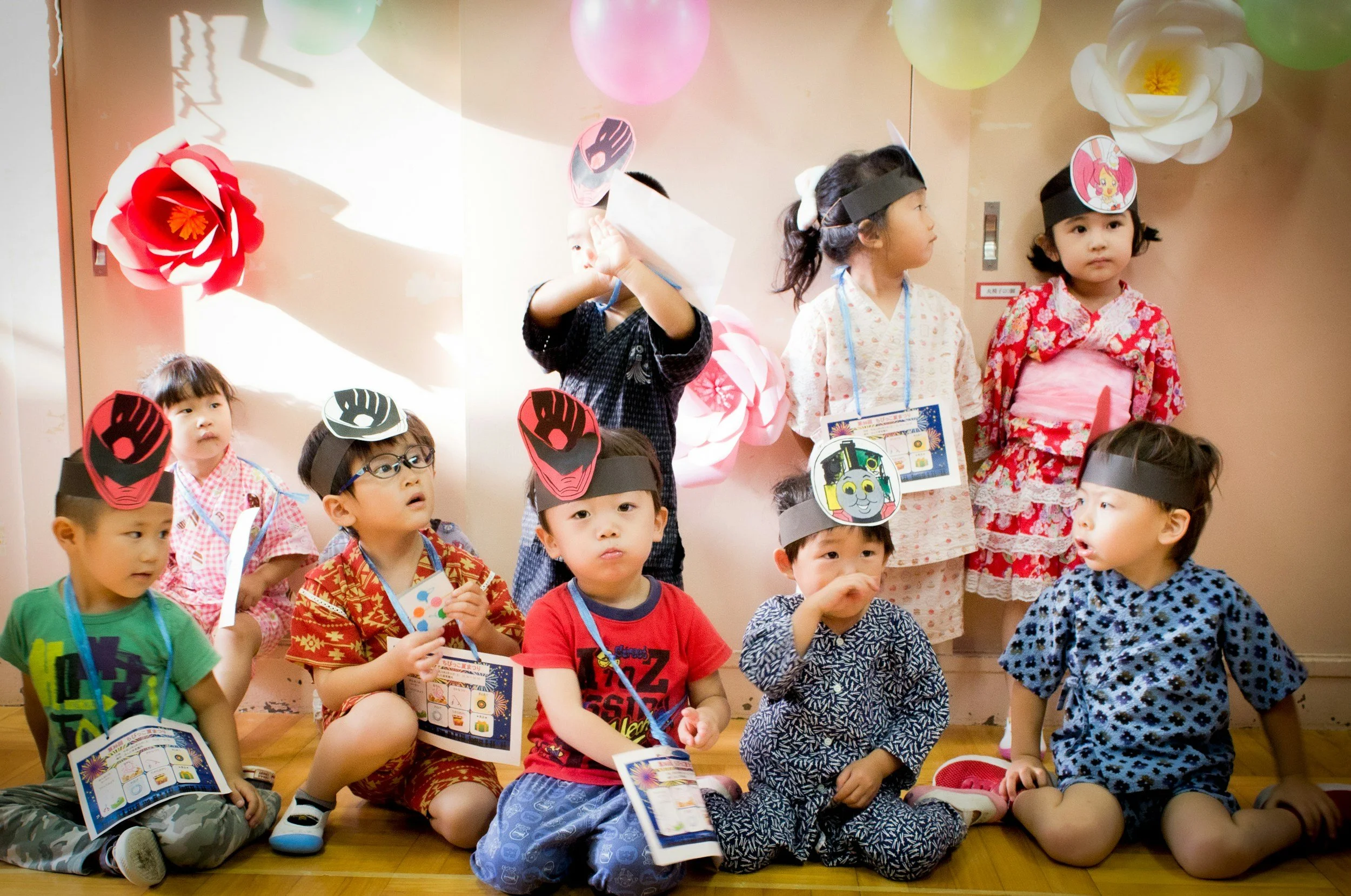 Group of young children wearing traditional Japanese yukatas and festive paper headbands, sitting on the floor wearing name tags and holding pamphlets, in a decorated room with paper flowers and colorful balloons.