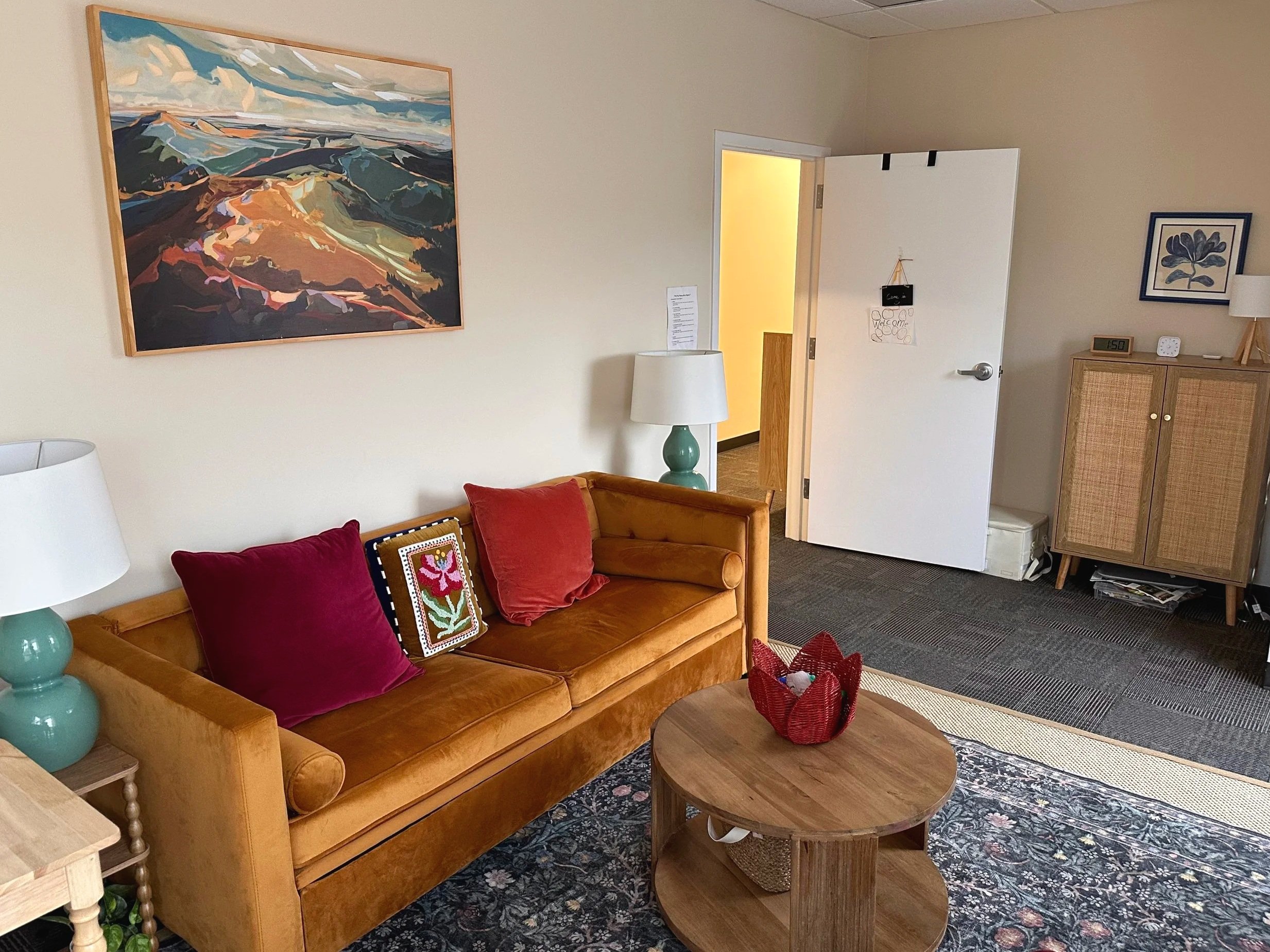 A cozy living room with a tan sofa adorned with red and purple pillows, a round wooden coffee table with a decorative red basket, a white lamp with a teal base, a large colorful landscape painting on the wall, and a doorway leading to a yellow-lit ro