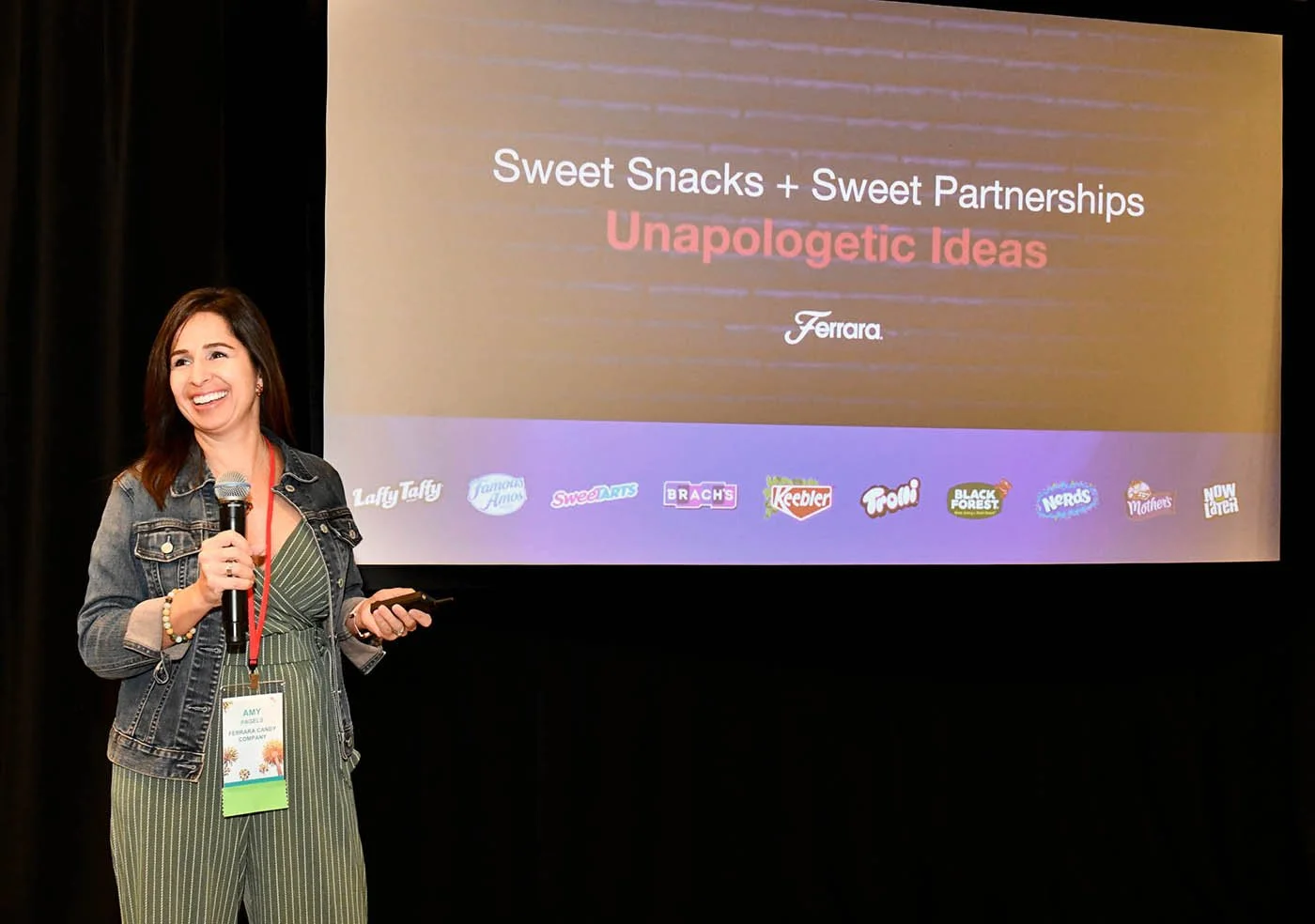 Woman smiling and holding a microphone and a remote control, standing in front of a presentation slide at a conference.