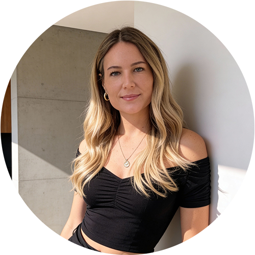 A young woman with long wavy blonde hair wearing a black off-shoulder top, standing against a background of concrete wall and white wall, with natural lighting.