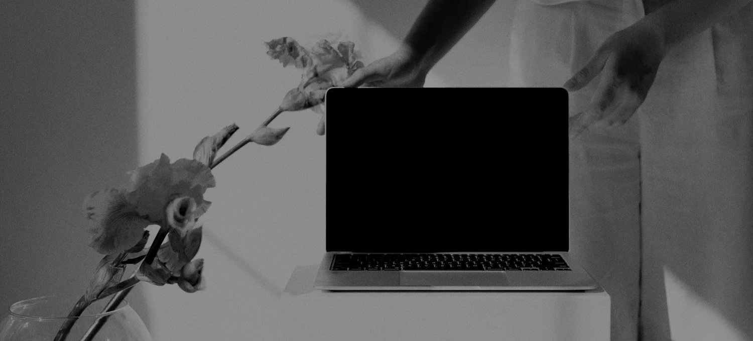 A person is arranging wilted flowers on a table next to a laptop with a black screen, in a grayscale photo.