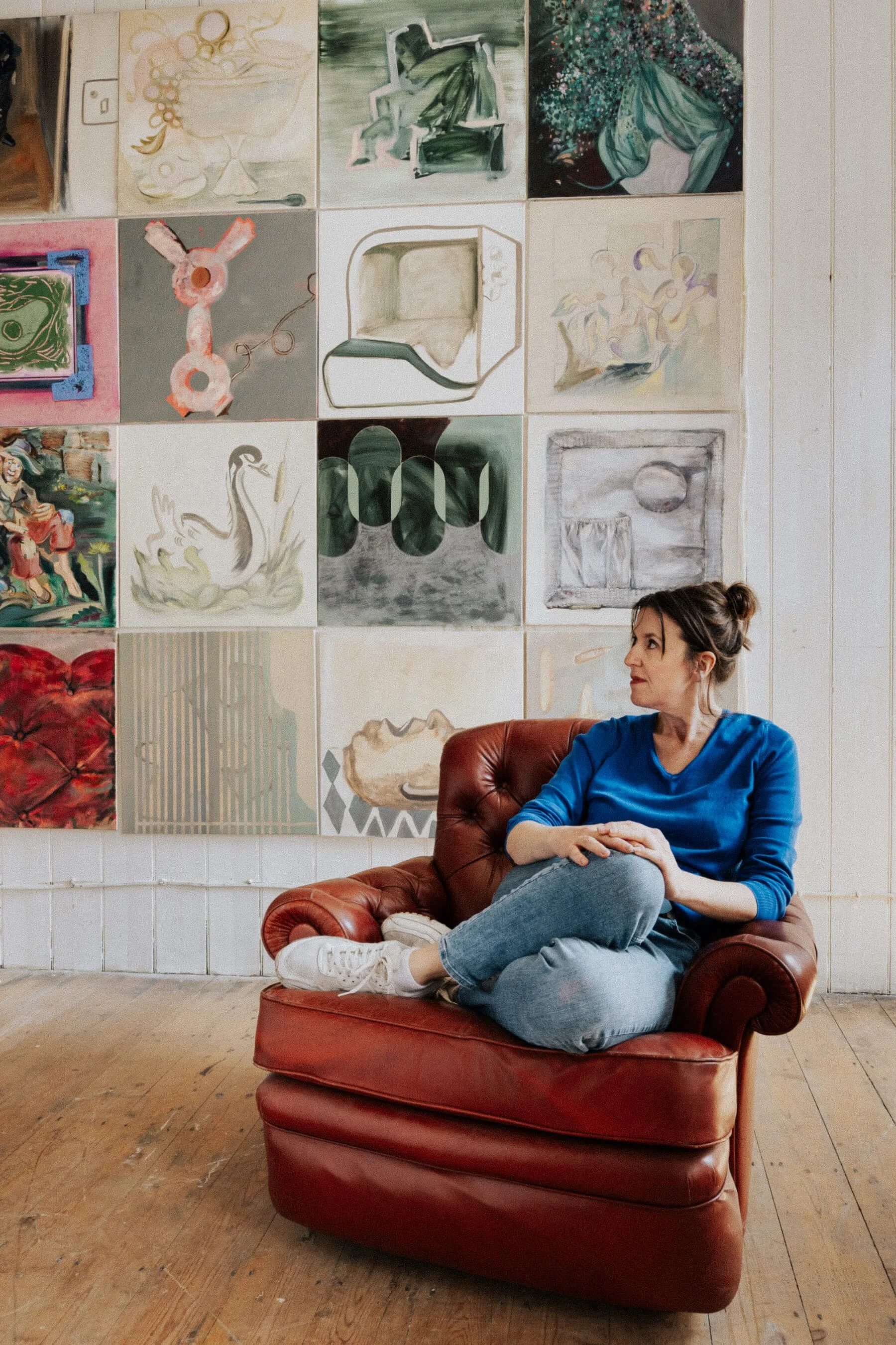 A woman in a blue shirt and jeans sitting on a brown leather armchair in an art gallery, with a colorful collage of paintings on the wall behind her.