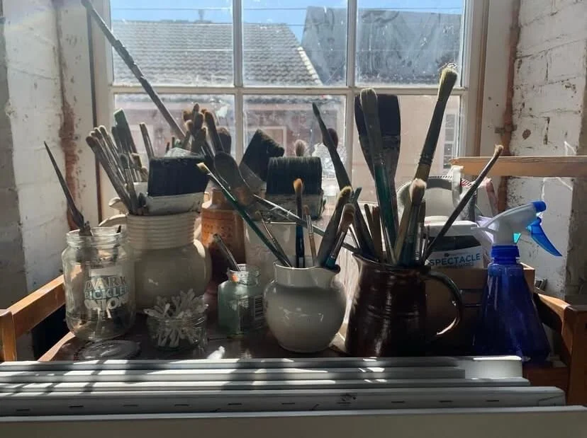 A collection of paint brushes and other art supplies on a table near a window in a bright art studio.