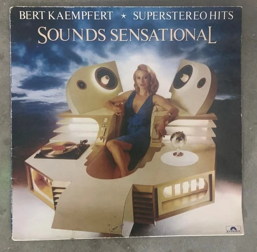 Album cover for Bert Kaempfert's 'Superstereo Hits: Sounds Sensational' featuring a woman sitting on a futuristic, rounded music studio with large speaker graphics against a dark, cloudy sky background.