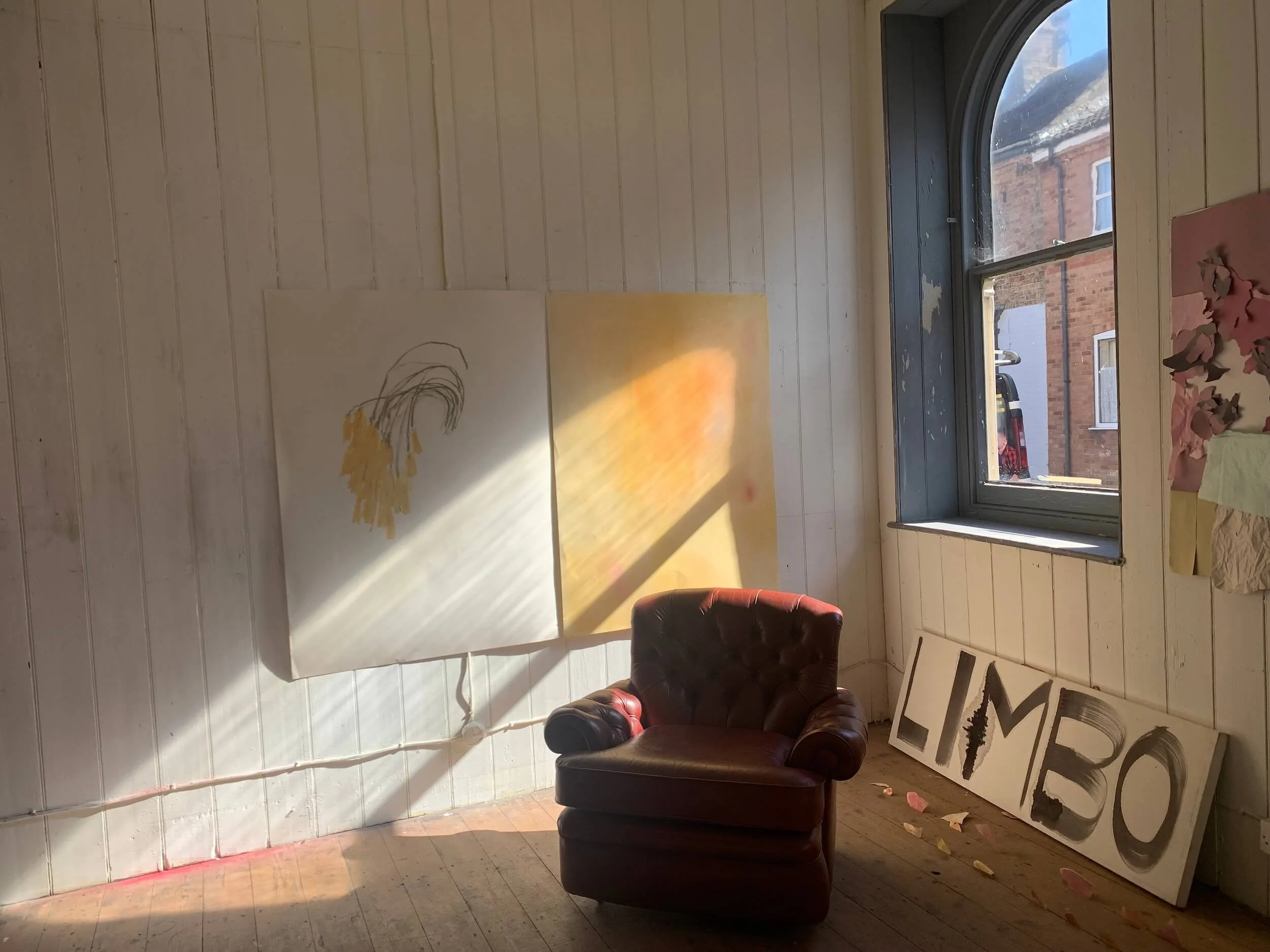 Sunlight coming through a window illuminates an art studio with a leather armchair, two paintings leaning against the wall, and a sign that says 'LIMBO' on the floor.