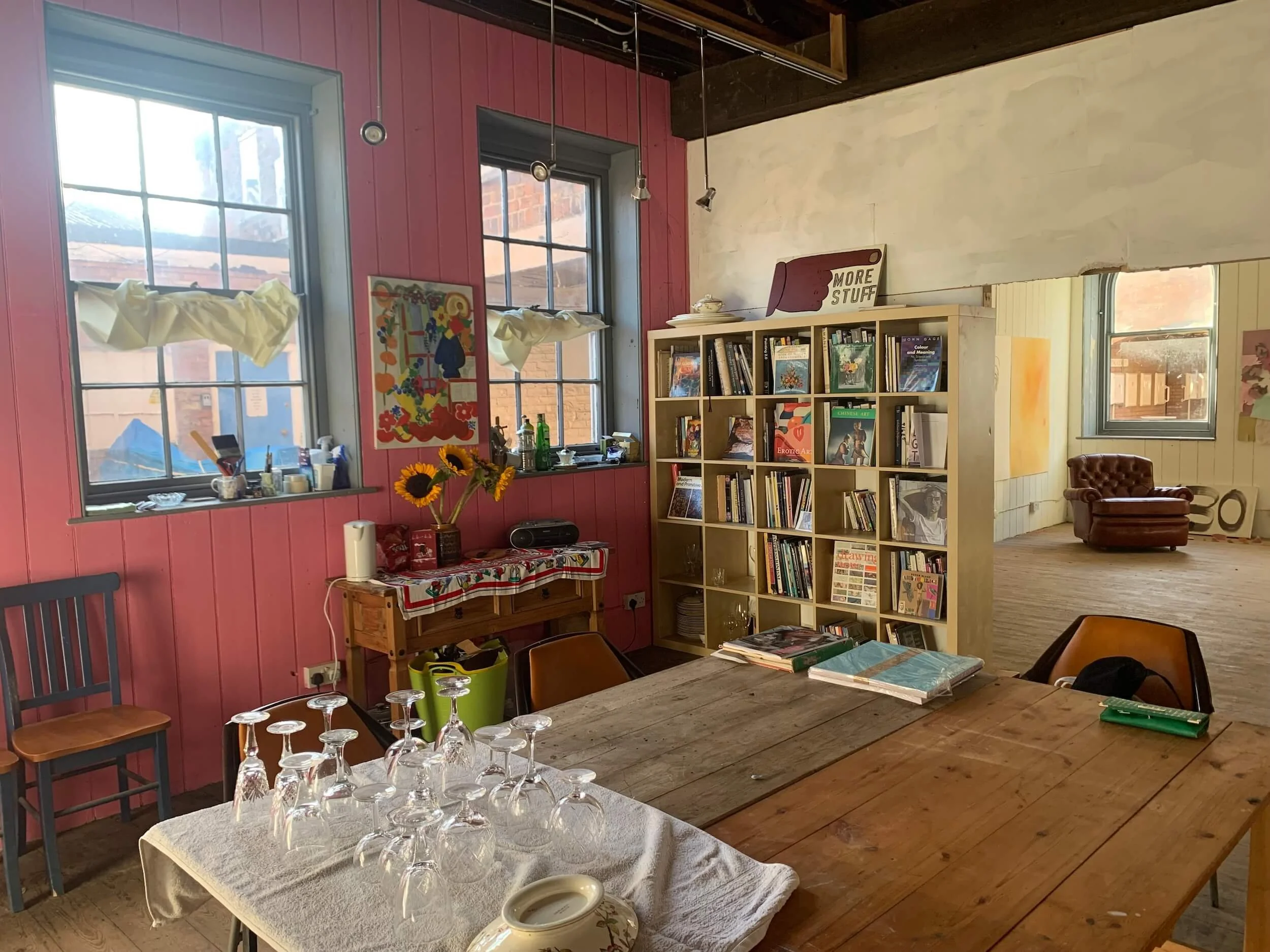 Interior of a room with pink and cream walls, two large windows, a wooden table with wine glasses, chairs, a bookshelf, and a brown leather armchair.