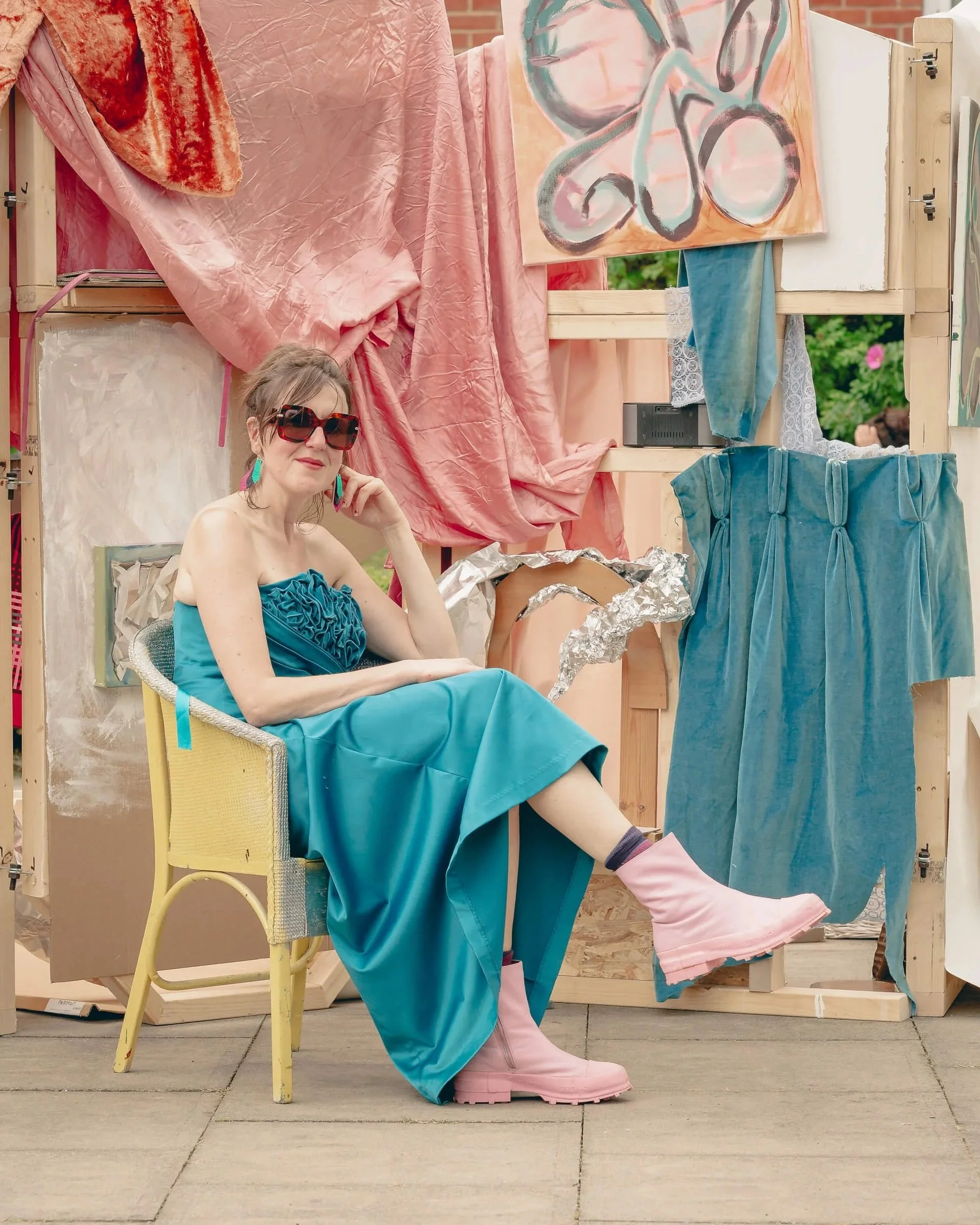 A woman sitting in a yellow chair wearing a teal dress, pink boots, large sunglasses, and earrings, with a creative art display background that includes fabric, paintings, and textured materials.