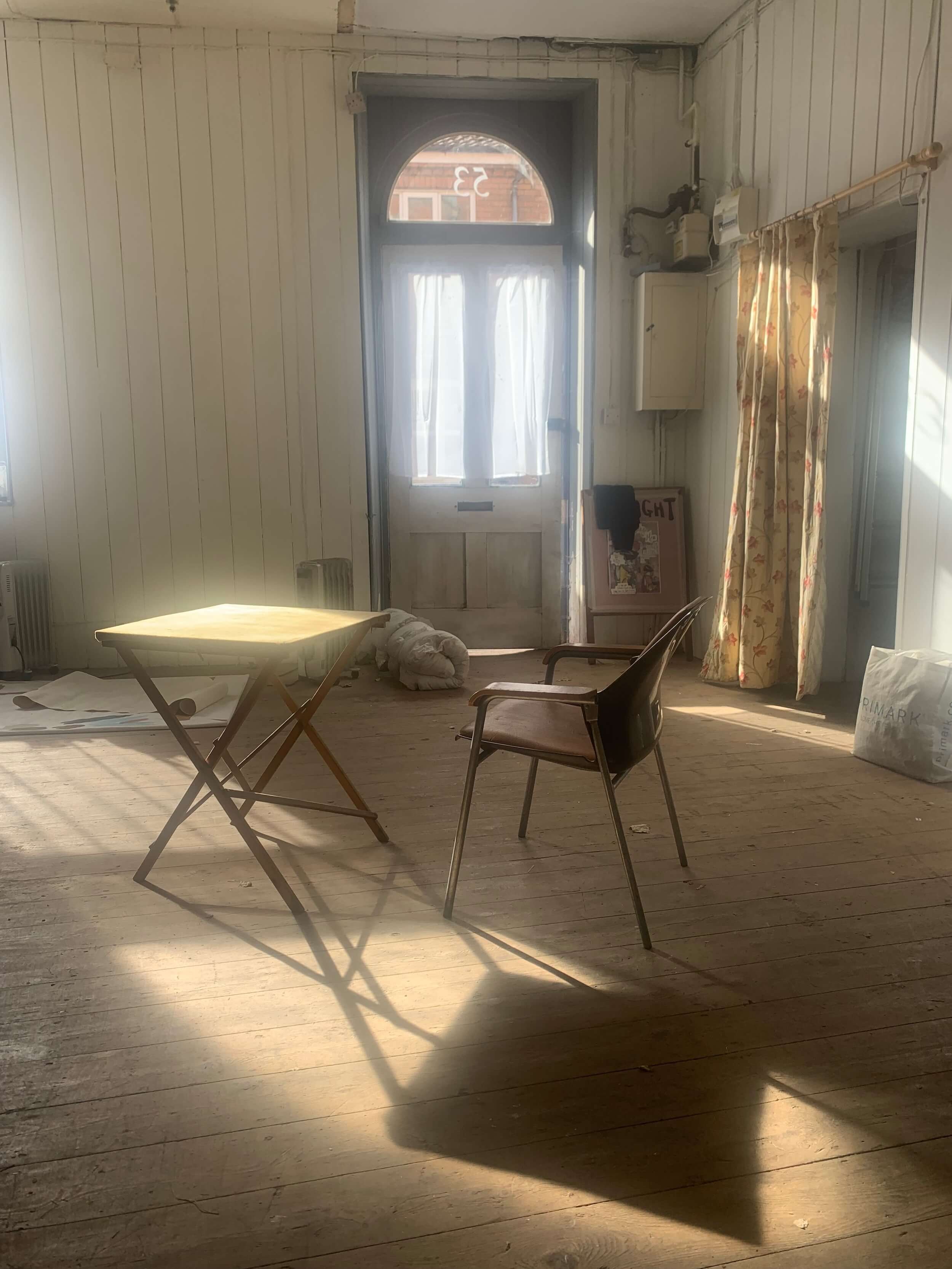 Sunlit room with wooden floor, a small table, a chair, curtains on the window, a door with a semi-circular window, and some items on the floor and shelves.