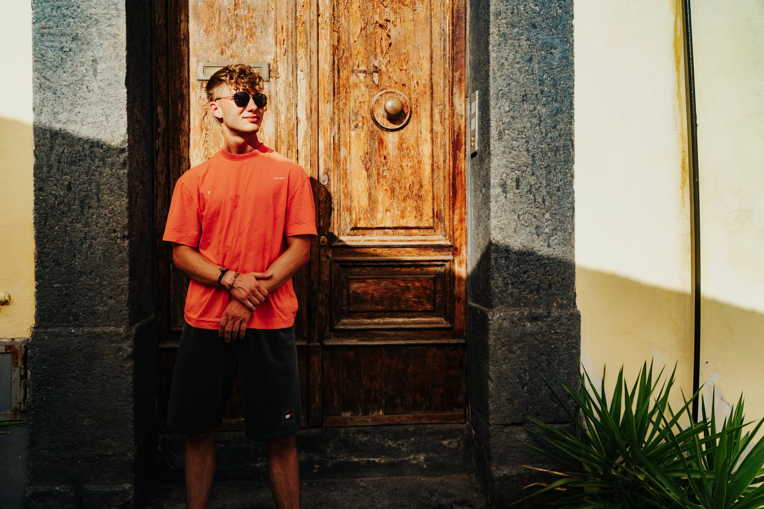 Junger Mann in Sonnenbrille und rotem T-Shirt steht vor einer Holztür und lächelt.
