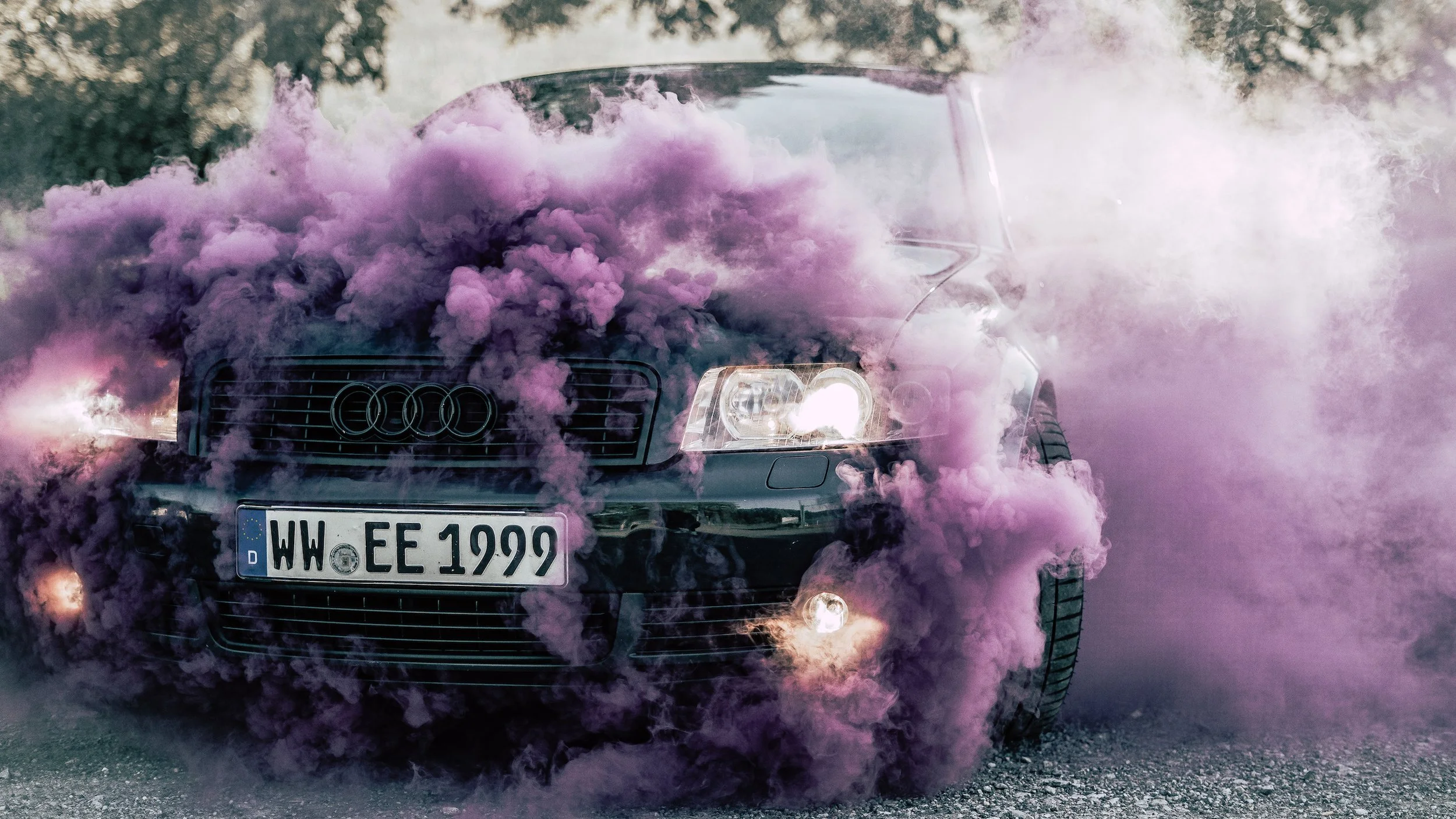 Ein schwarzer Audi-Wagen mit rosa Rauch und Nebel umhüllt auf einer offenen Fläche, mit sichtbarer Mercedes-Nummernschild."