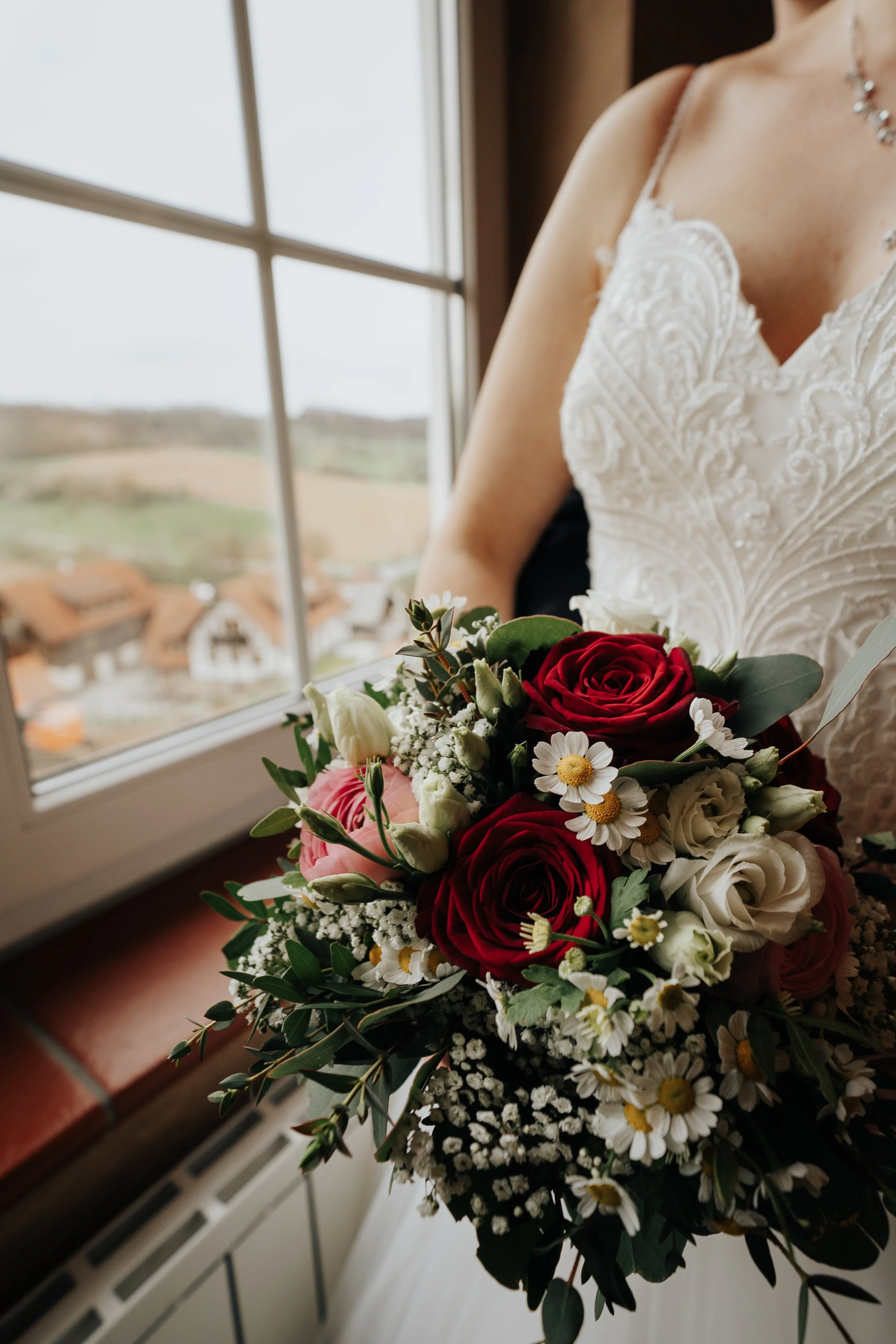 Braut hält einen Blumenstrauß mit roten, weißen und rosa Blumen vor einem Fenster mit Blick auf eine ländliche Landschaft.