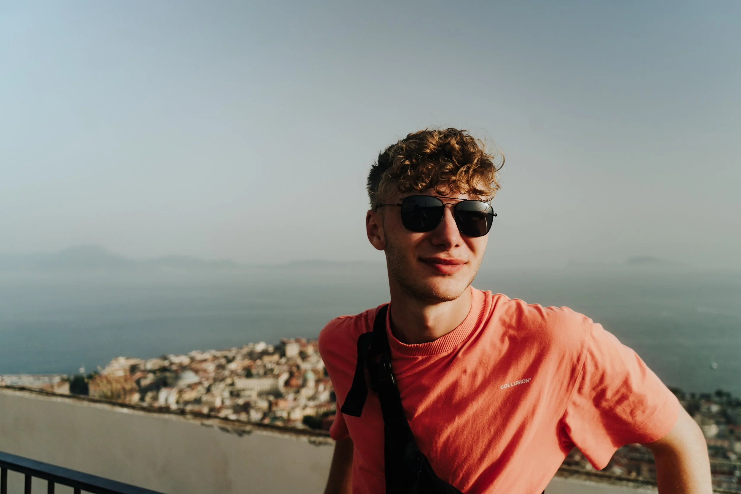 Junge Mann mit Sonnenbrille auf einer Terrasse mit Blick auf das Meer und eine Stadt im Hintergrund, bei Sonnenuntergang.
