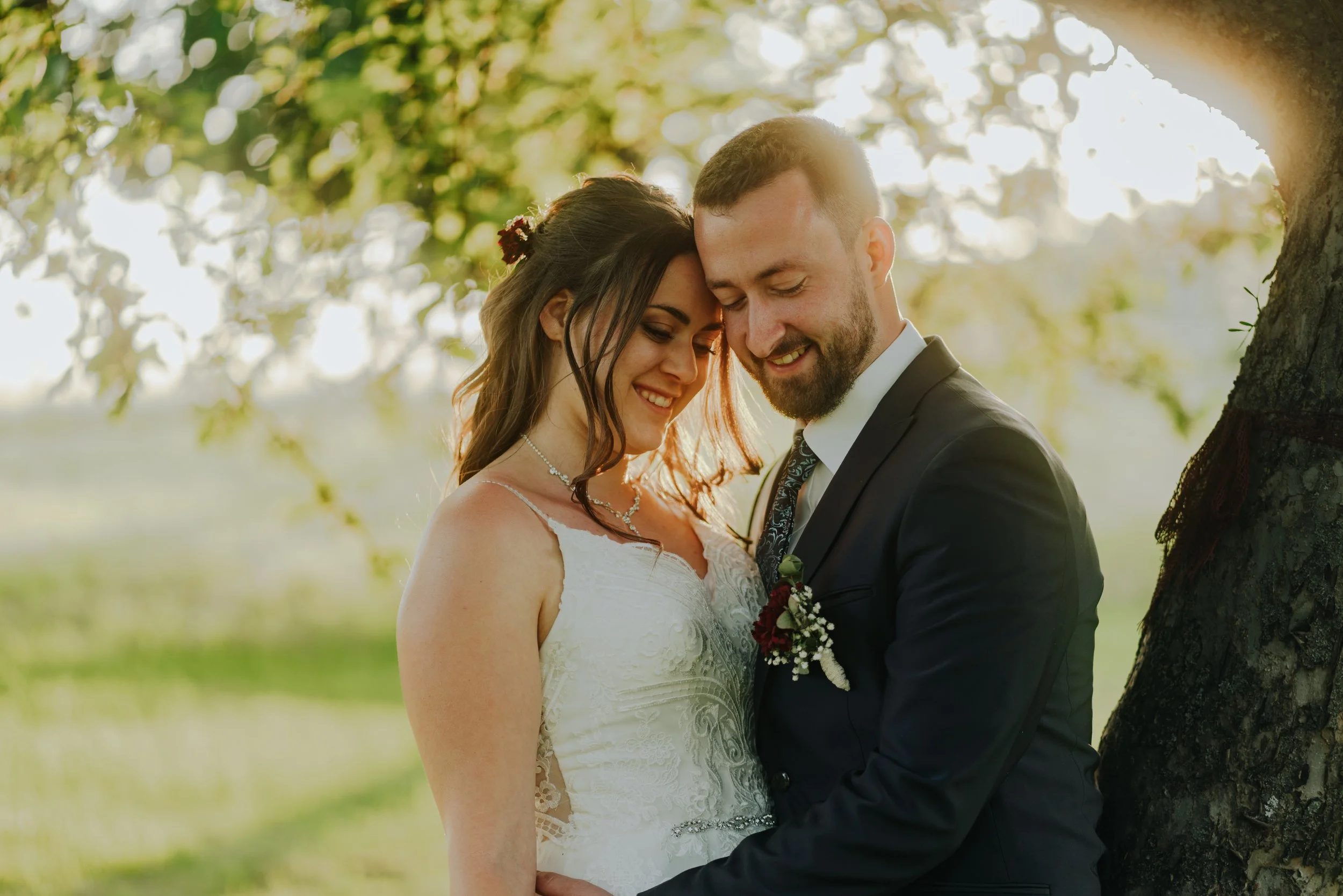 Ein Brautpaar steht unter einem Baum, umarmt sich und lächelt bei Sonnenuntergang im Freien.