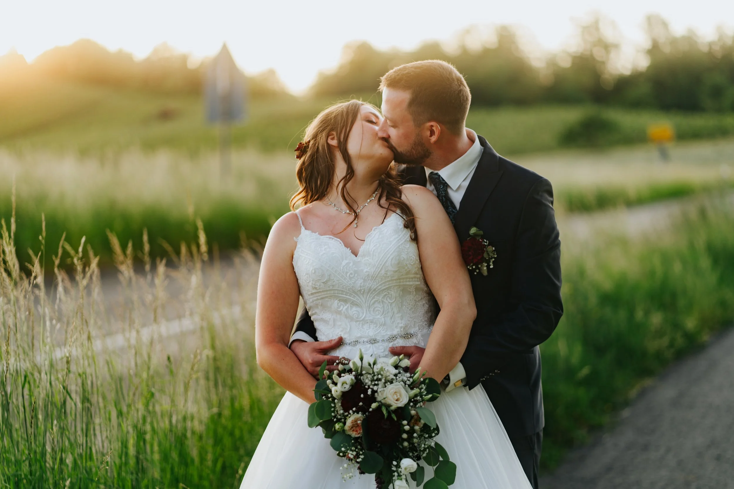 Ein frisch verheiratetes Paar küsst sich im Sonnenuntergang in einer grünen Landschaft, die Braut trägt ein weißes Hochzeitskleid und hält einen Blumenstrauß, der Bräutigam trägt einen dunklen Anzug.