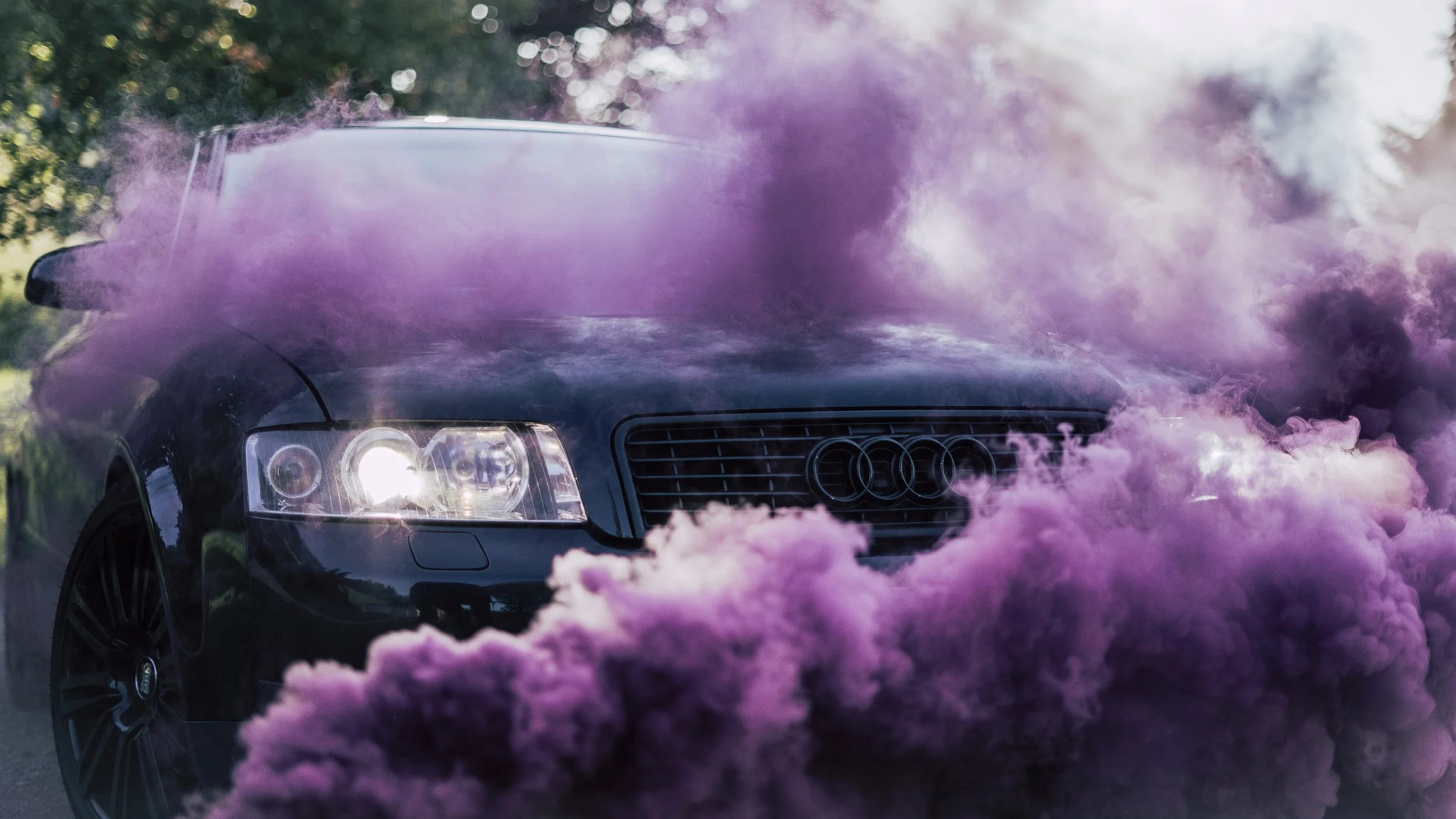 Ein schwarzer Audi-Auto, umgeben von violettem Rauch oder Nebel, im Freien bei Sonnenlicht.