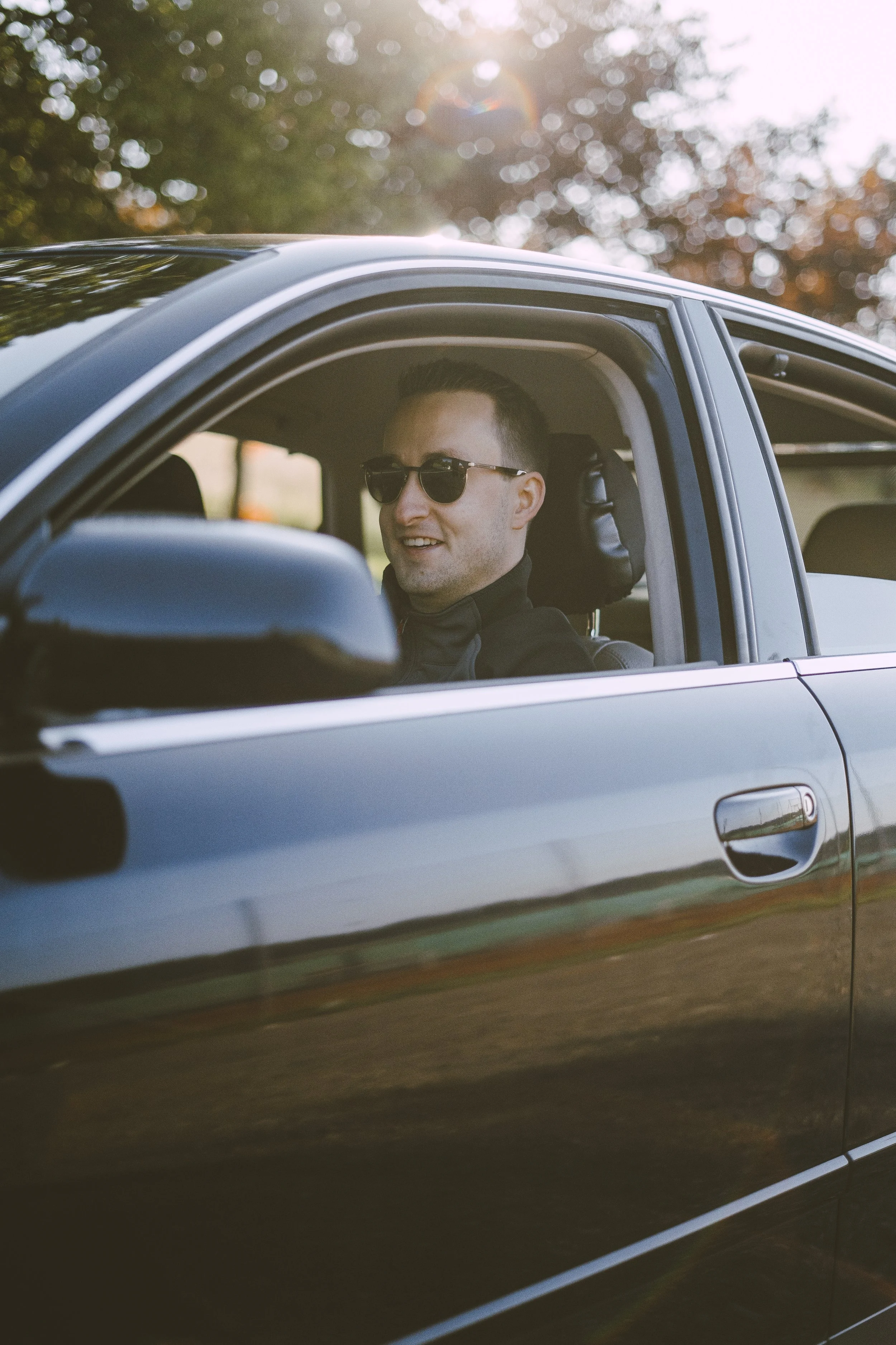 Ein Mann mit Sonnenbrille sitzt lächelnd in einem schwarzen Auto bei Sonnenuntergang, im Hintergrund Bäume mit herbstlichen Blättern.
