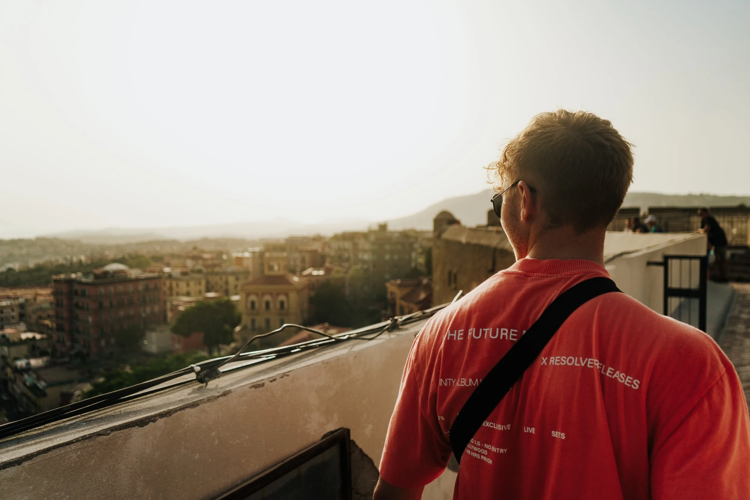 Junger Mann mit Sonnenbrille und rotem T-Shirt schaut von einer Dachterrasse auf eine Stadt bei Sonnenuntergang.