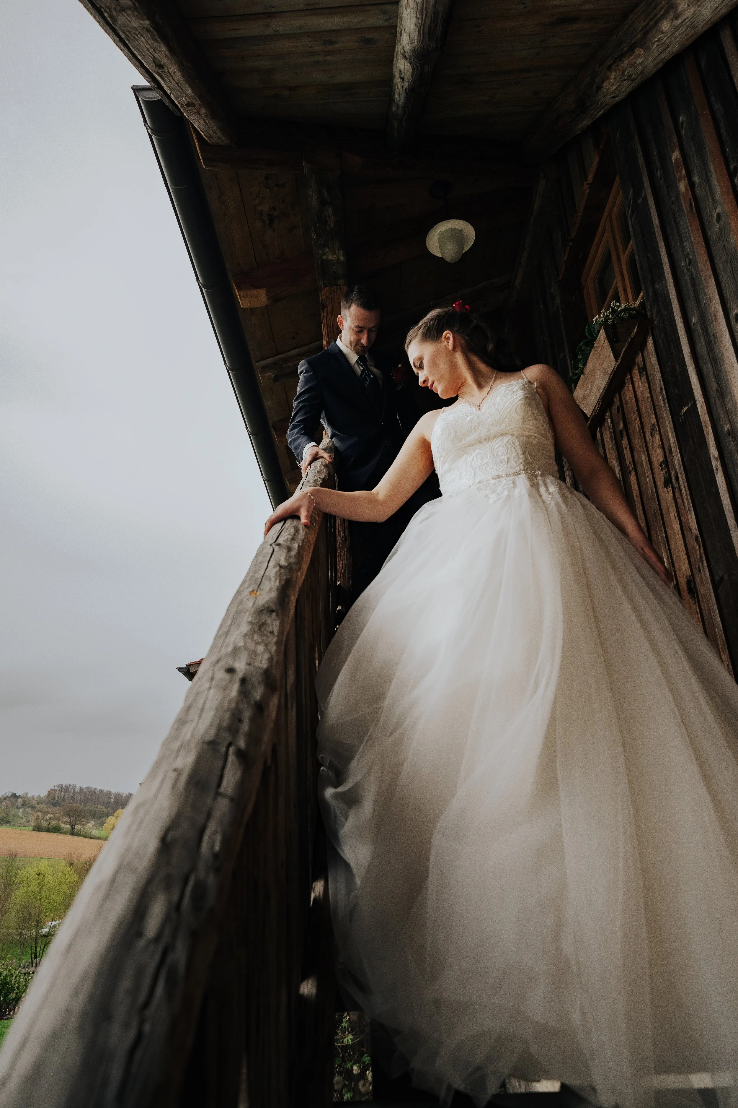 Ein Brautpaar auf einem Holzbalkon, die Frau trägt ein weißes Brautkleid, der Mann trägt einen Anzug, im Hintergrund eine ländliche Landschaft bei leicht bewölktem Himmel.