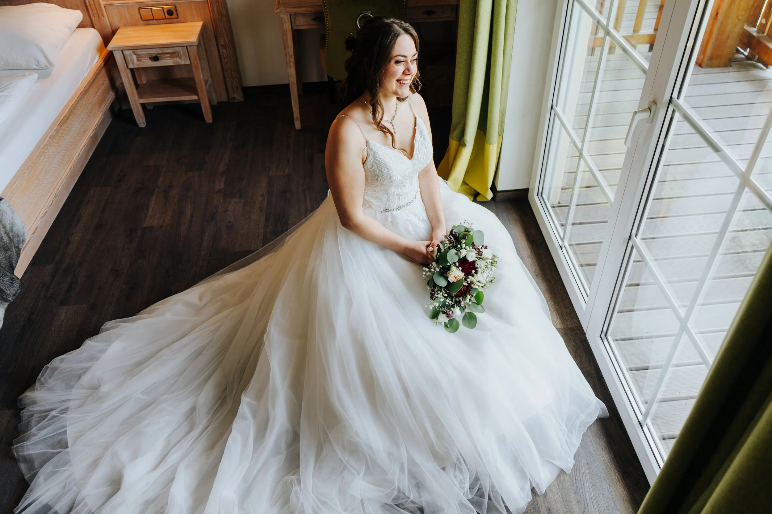 Braut mit weißem Hochzeitskleid sitzt auf dem Boden, hält einen Blumenstrauß und lächelt, während sie auf einem Holzfußboden vor einem großen Fenster sitzt.