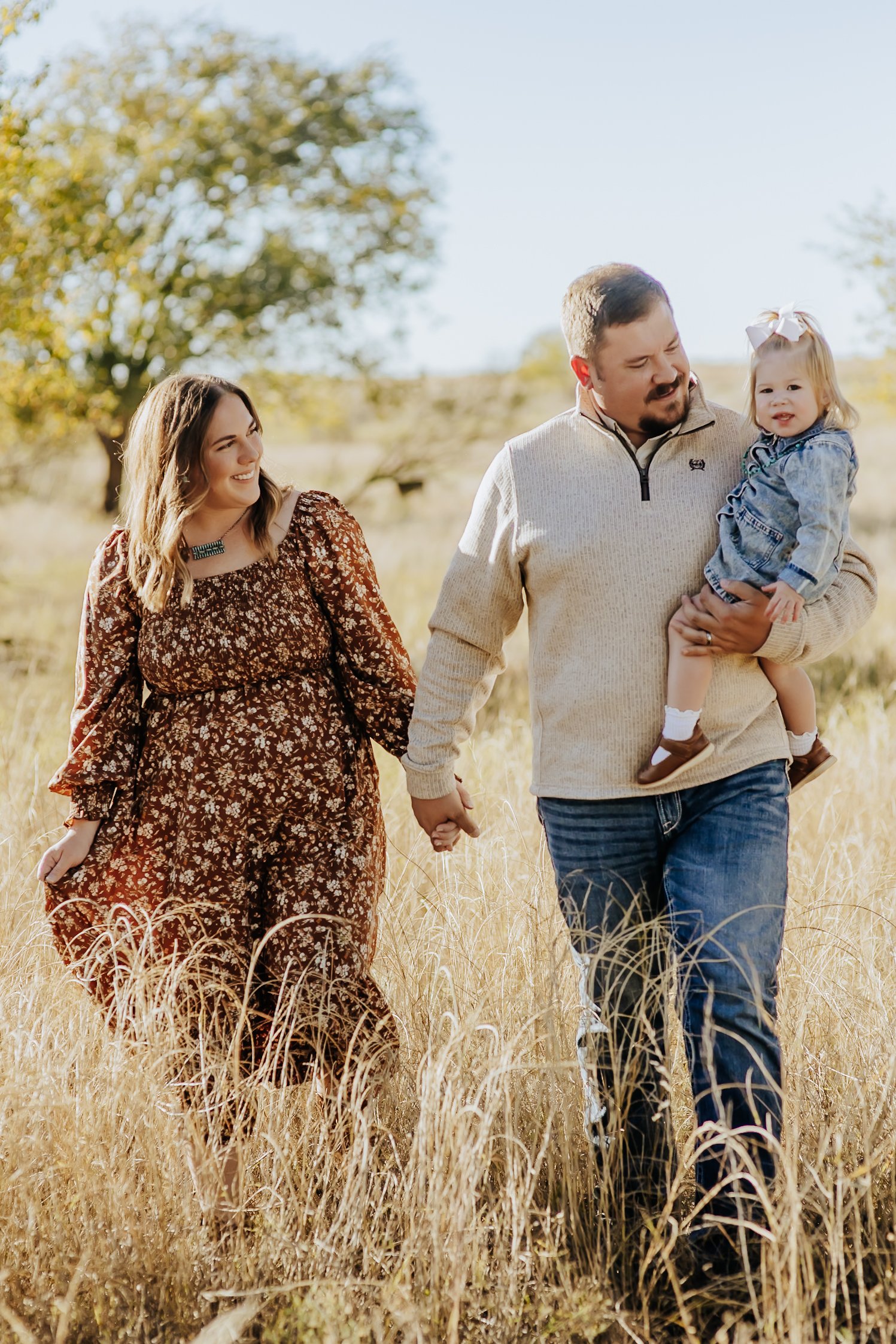 A family of three walking hand-in-hand through a golden field of tall grass on a sunny day, with trees in the background.