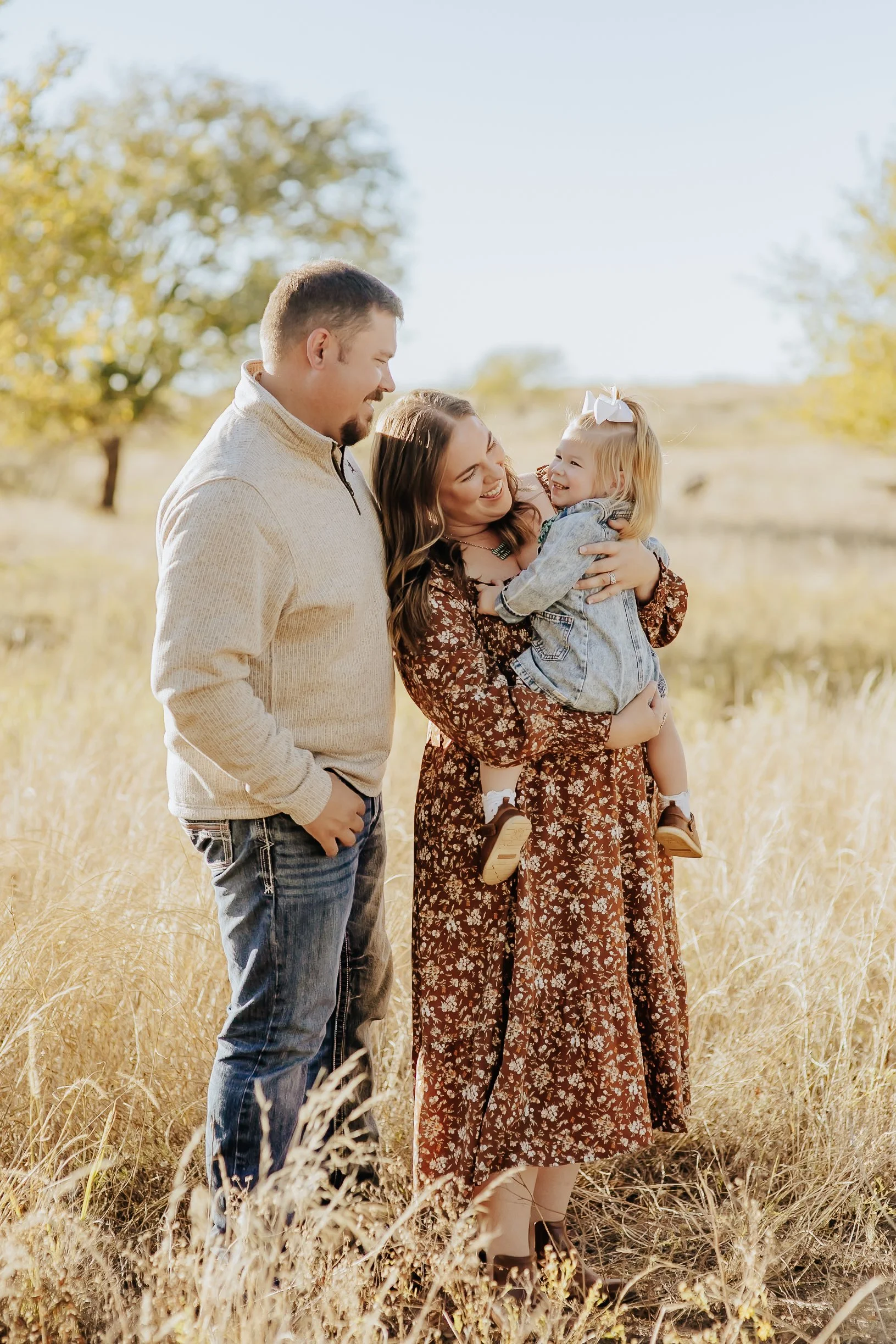 A family of three standing in a field of tall grass, smiling and looking at each other, with trees and a clear sky in the background.