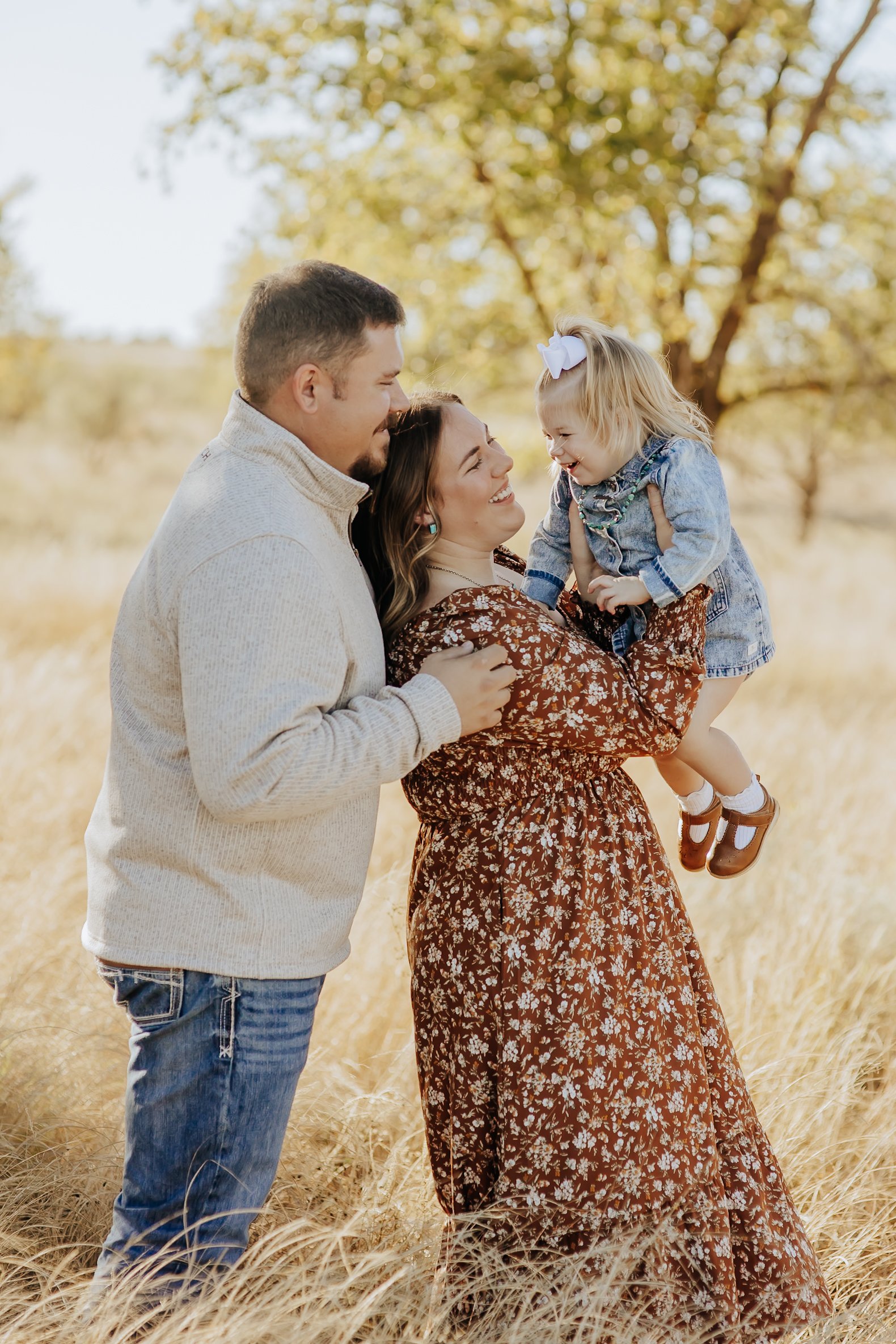 A family of three outdoors in a field with tall grass, smiling and playing together. The woman is holding a young girl, and a man is touching her shoulder, all engaged in a joyful moment.