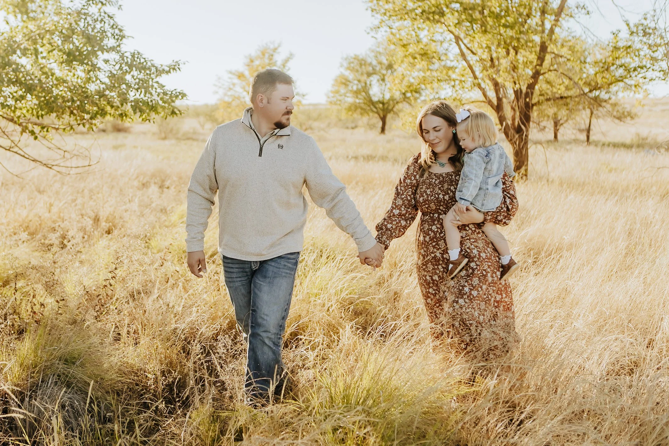 A family of three walking hand in hand through a field of tall yellow grass during autumn, with trees and a clear sky in the background.