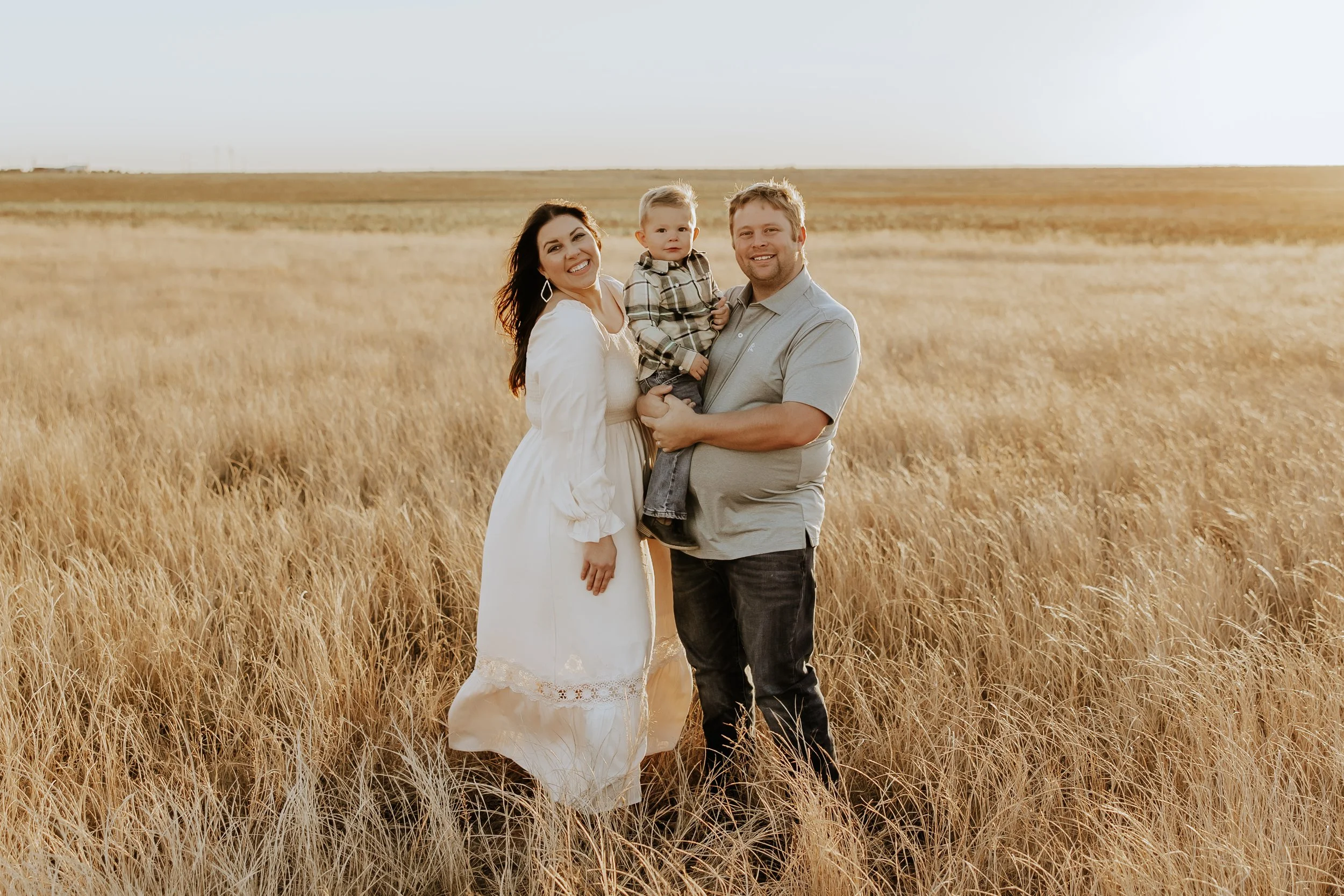A family of three standing in a golden field during sunset, smiling at the camera. The woman is wearing a white dress, the man is in a light gray shirt and jeans, and the child is in a plaid shirt and jeans.