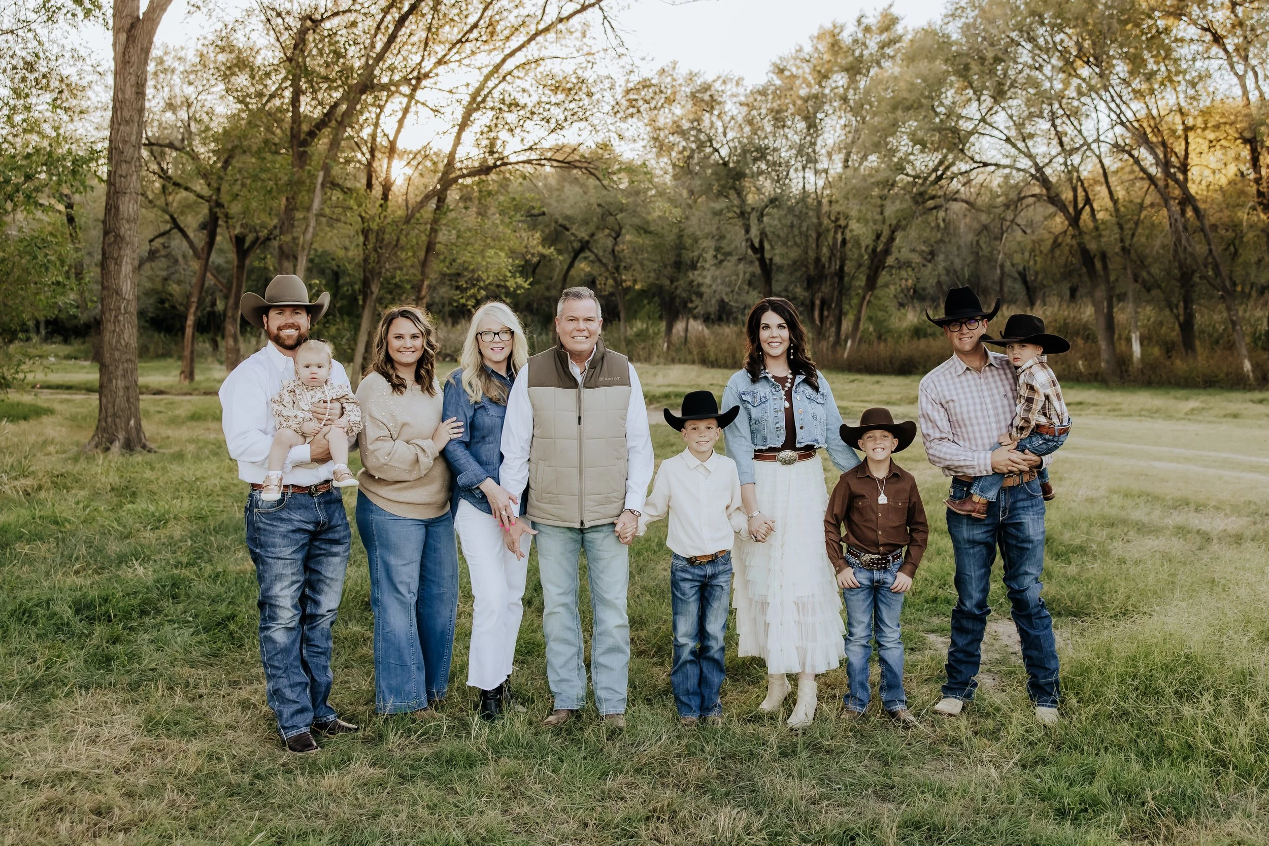 Family of ten posing outdoors in a grassy park during sunset, wearing casual and western-style clothing, including cowboy hats and boots, with trees in the background.