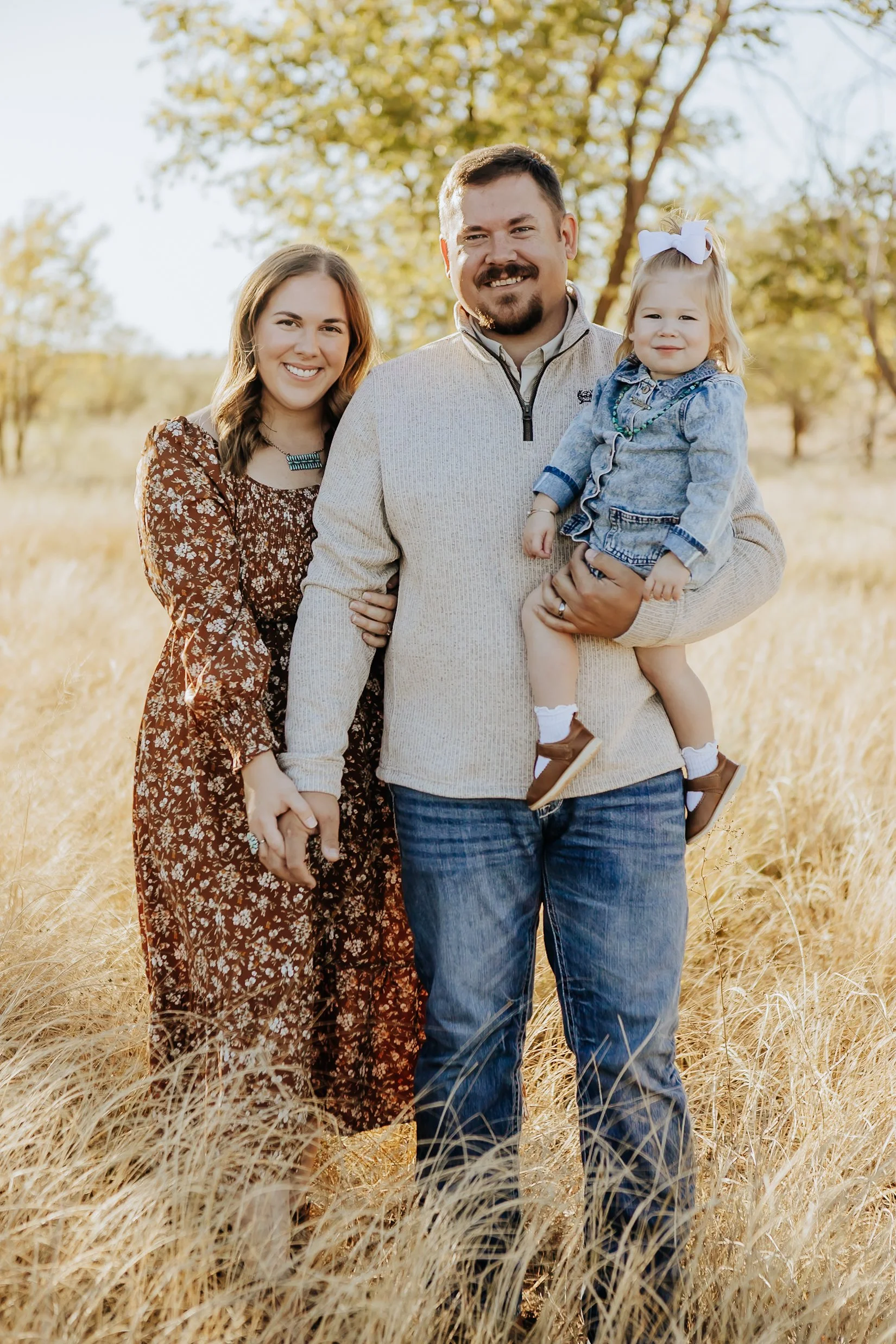 A family of three standing in tall grass outdoors during autumn, with trees in the background. The man is holding a little girl, and a woman stands beside them, all smiling at the camera.