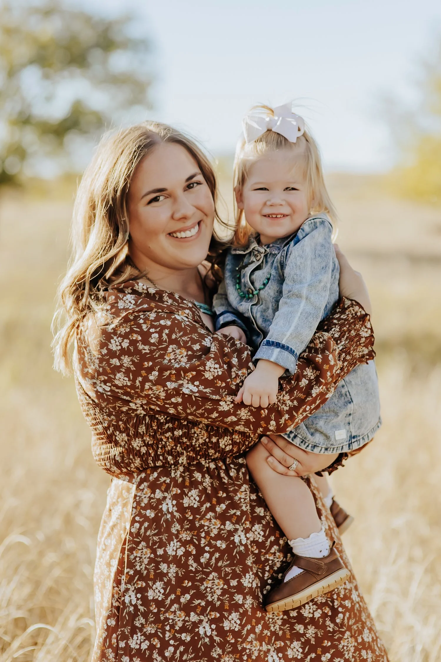 A smiling woman holding a young girl with a white bow in her hair outdoors in a field, both wearing casual clothing.