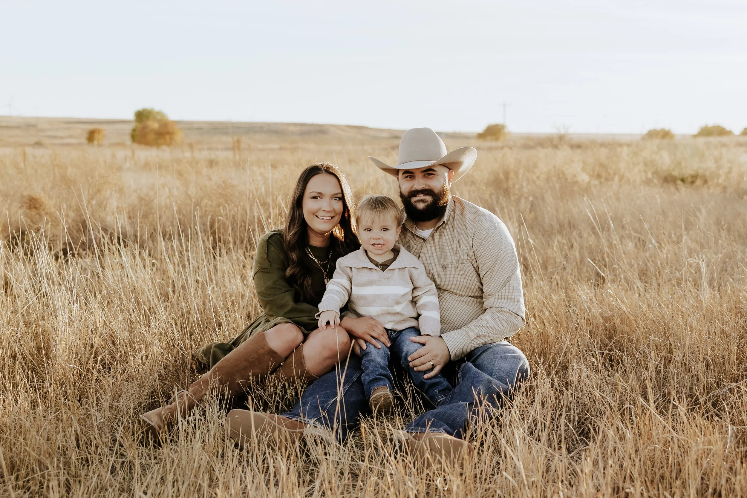 A family of three sitting in a field of tall, golden grass with a clear sky in the background. The woman has long brown hair, the man is wearing a cowboy hat and has a beard, and the young boy is sitting between them.