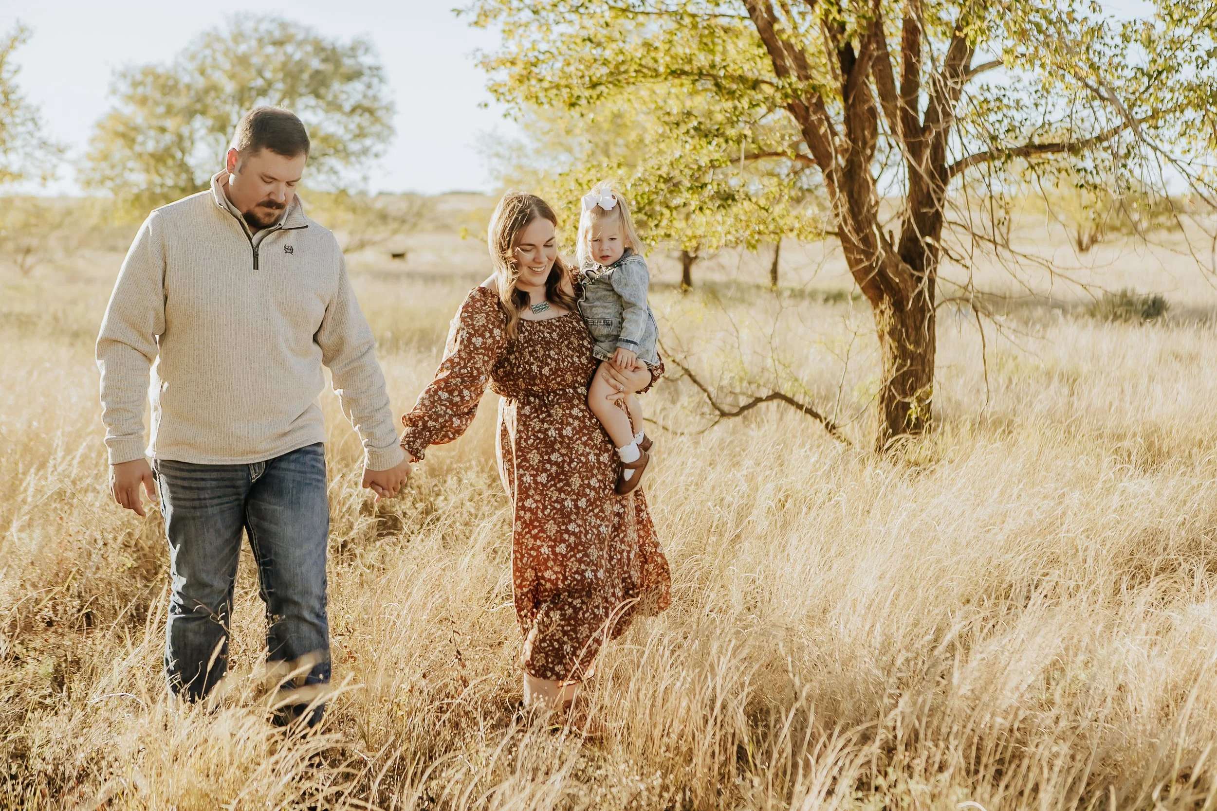 A family of three walking hand in hand through a grassy field during autumn, with a woman carrying a young girl and a man holding her hand, all smiling.