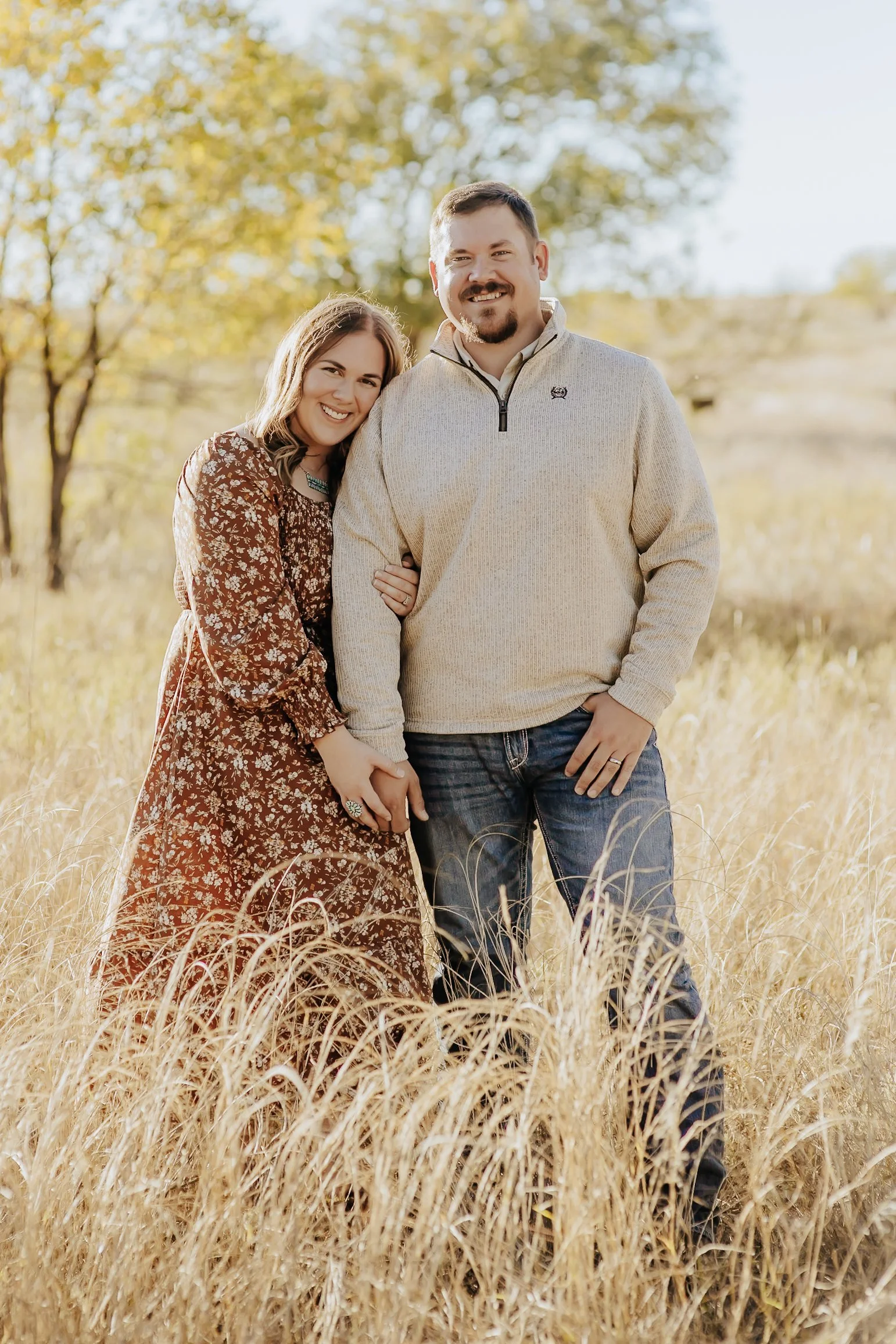 A couple standing close in a field of tall, golden grass with trees in the background, smiling and holding hands.