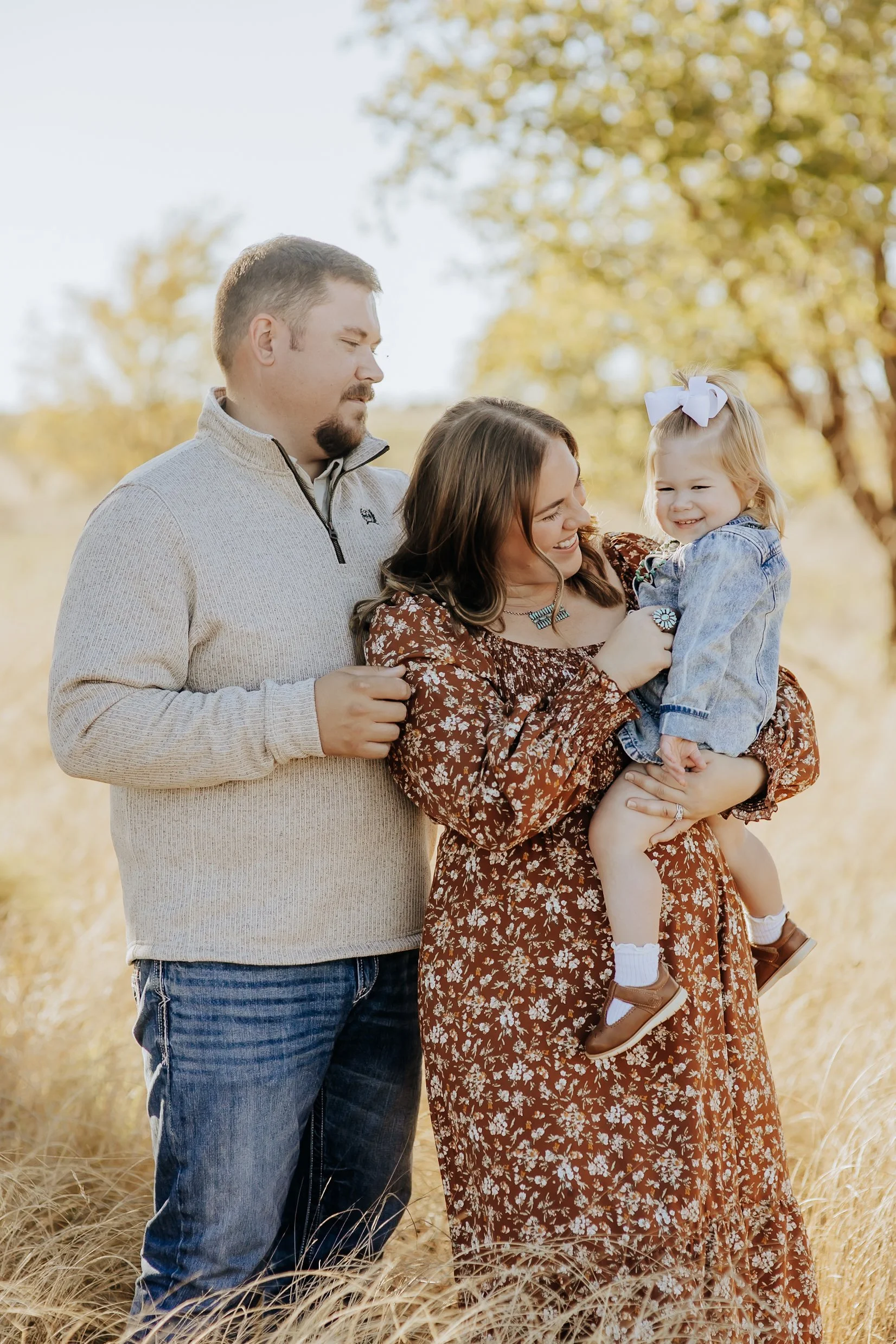 Family of three outdoors in a field during autumn, with a man, woman, and young girl. The woman is holding the girl, who has a big white bow in her hair. The scene is warmly lit.