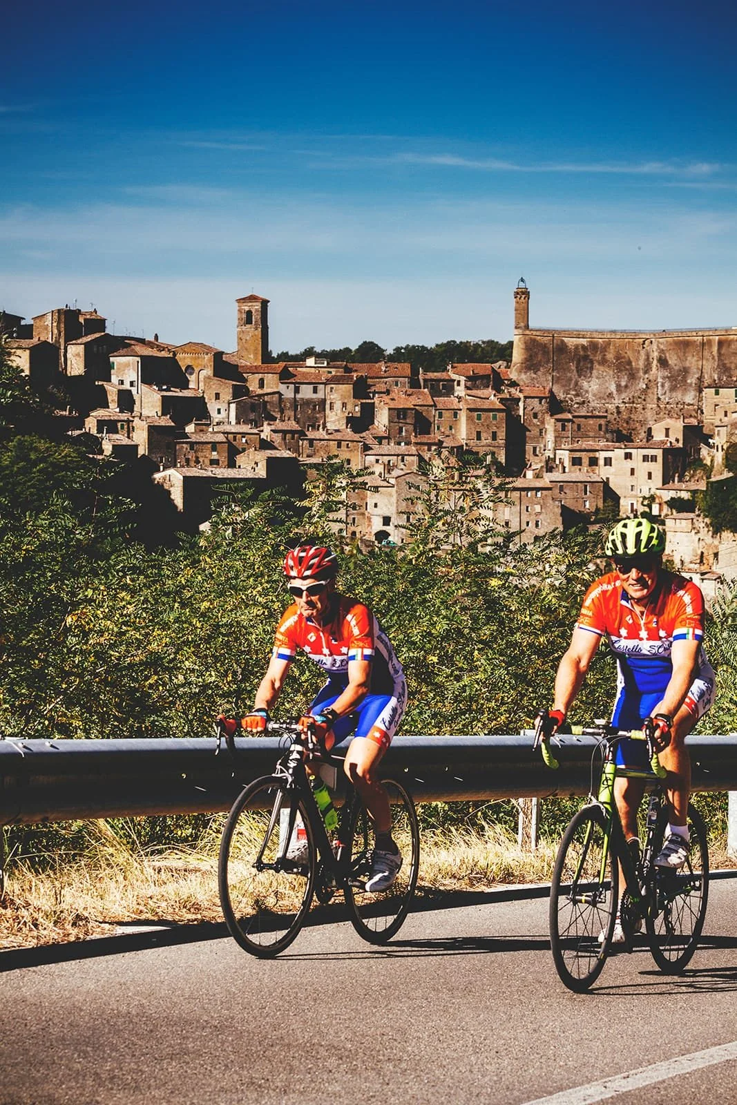 Two people cycling on a road with a traditional italian village in the background.