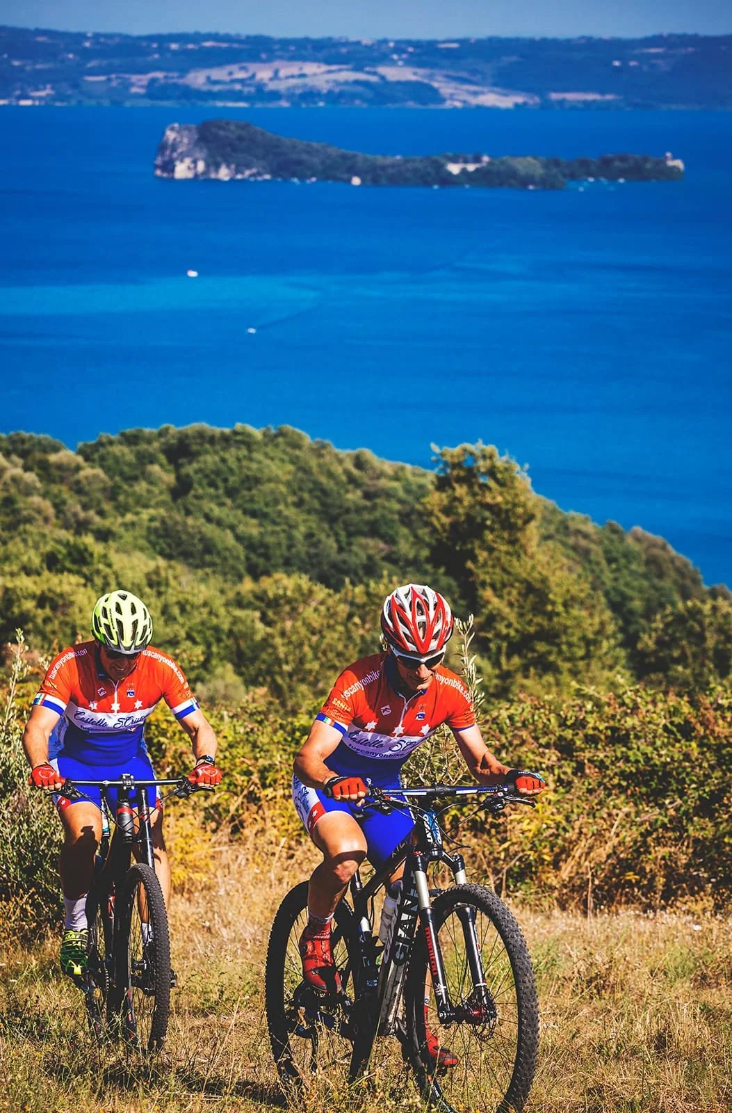Two people cycling on a trail with a blue lake in the background.