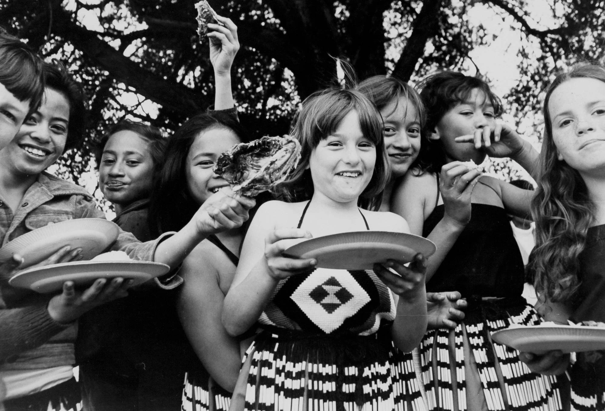 Black and white photo feaeturing Māori girls in piupiu holding a plate and passing food/kai